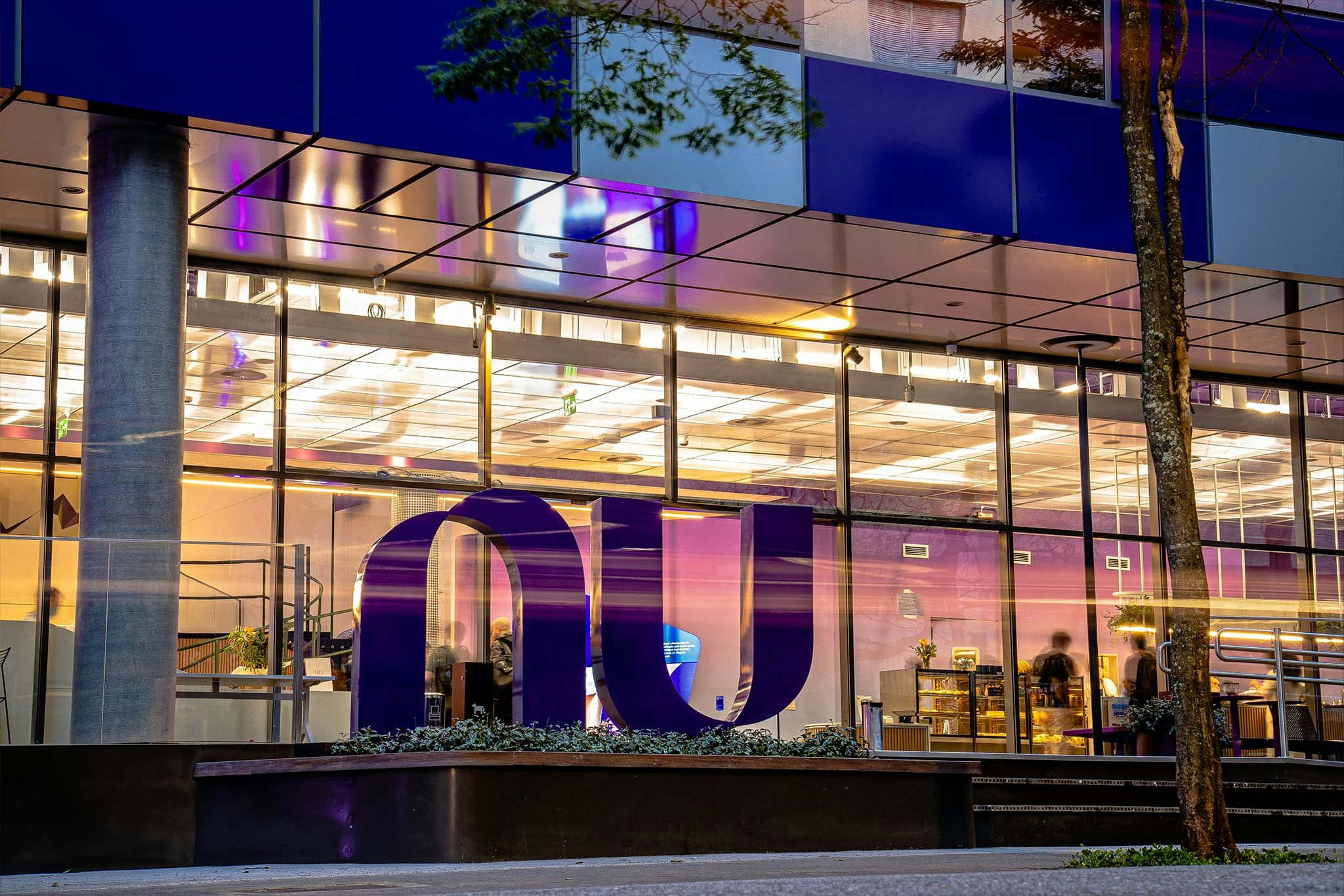 Logo roxo do Nubank em um pátio arborizado, com a fachada do escritório ao fundo.