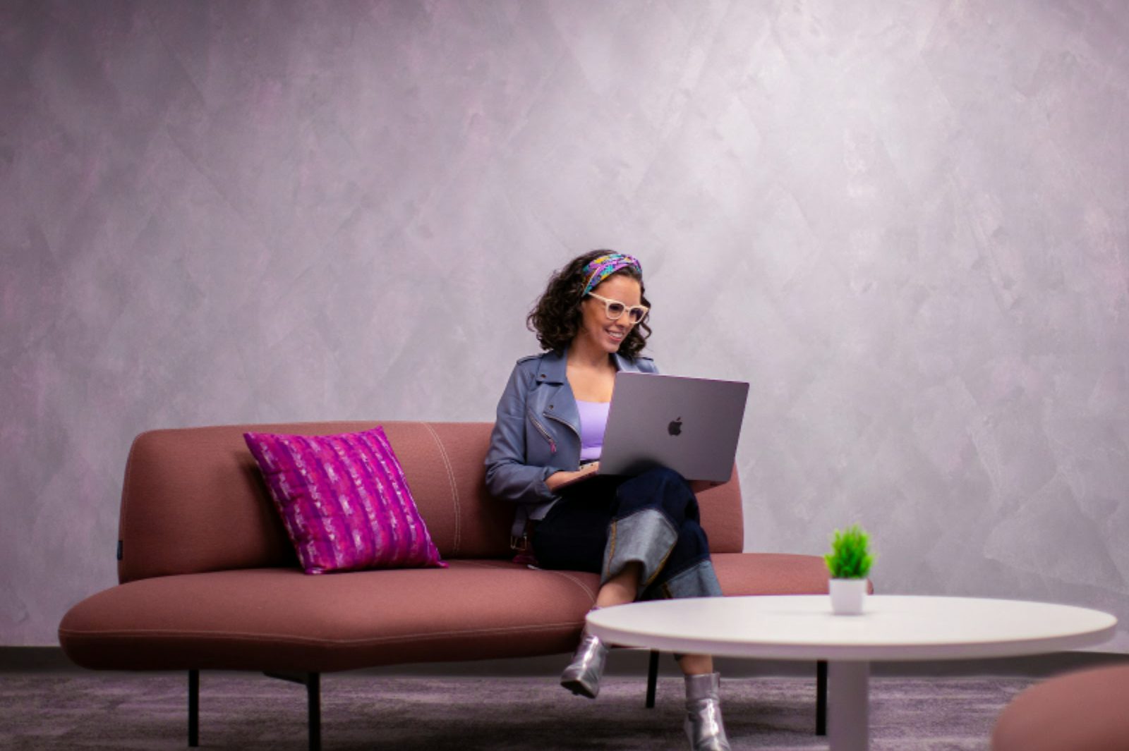 Mujer sonriente con gafas trabajando en una computadora portátil sentada en un moderno sofá rosa.