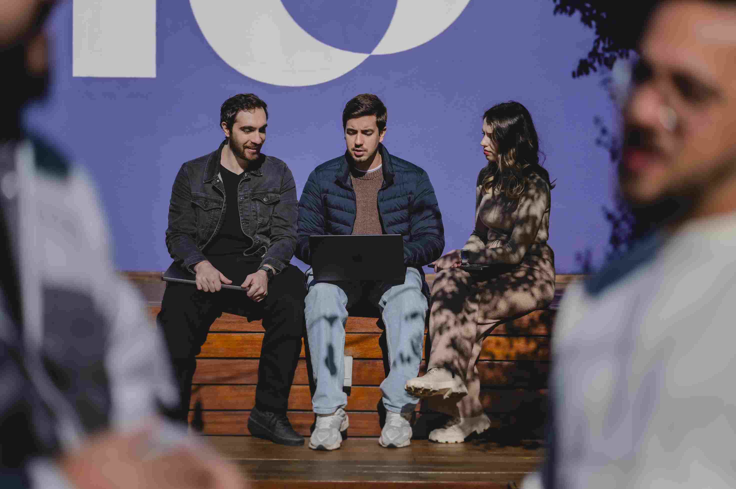 Three people talking on a wooden bench, one of them with a laptop, with a lilac wall and the Nubank symbol in white in the background.