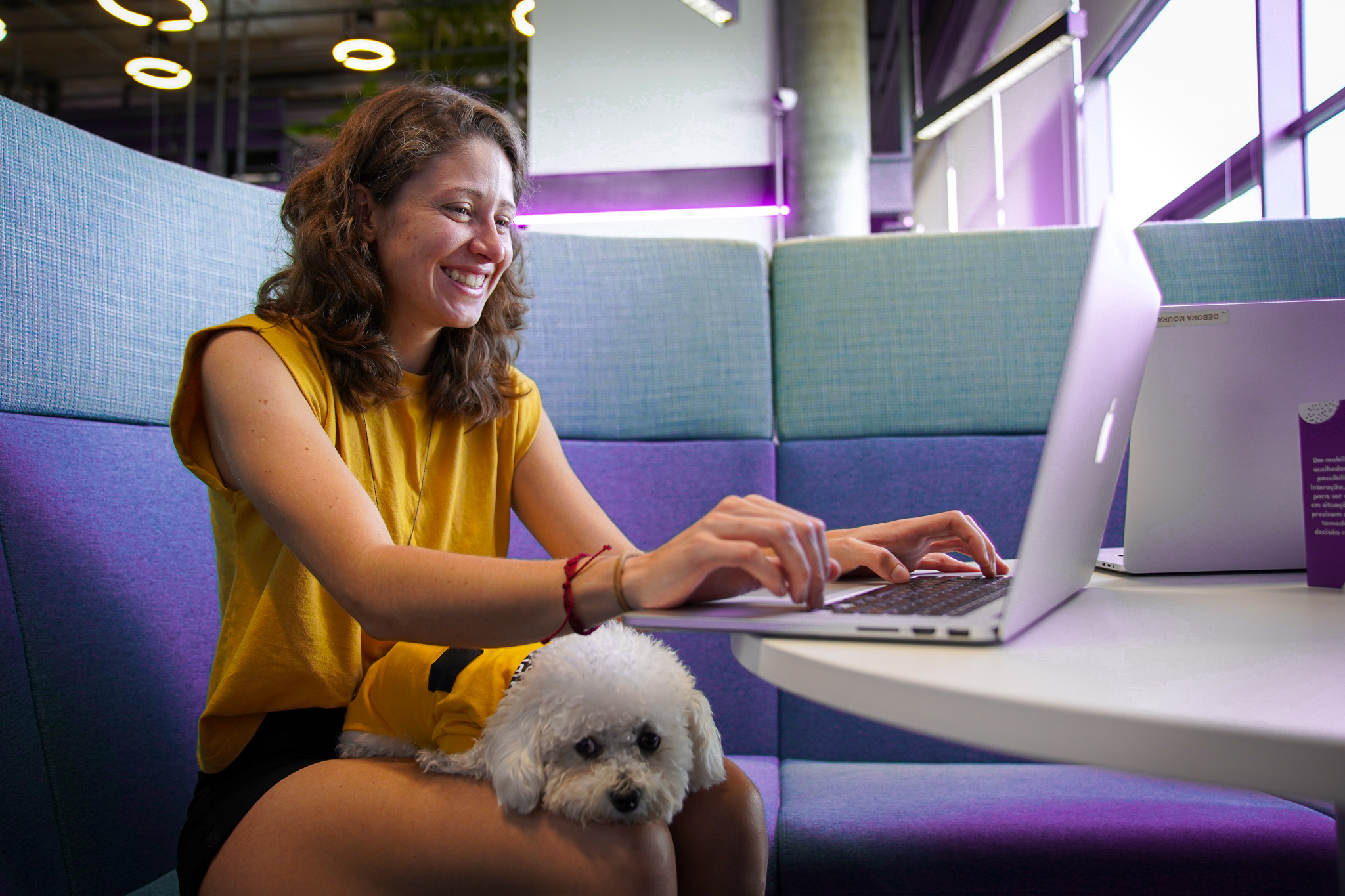 A woman in a yellow dress types on a laptop at an office lounge table, with a small white dog beside her.
