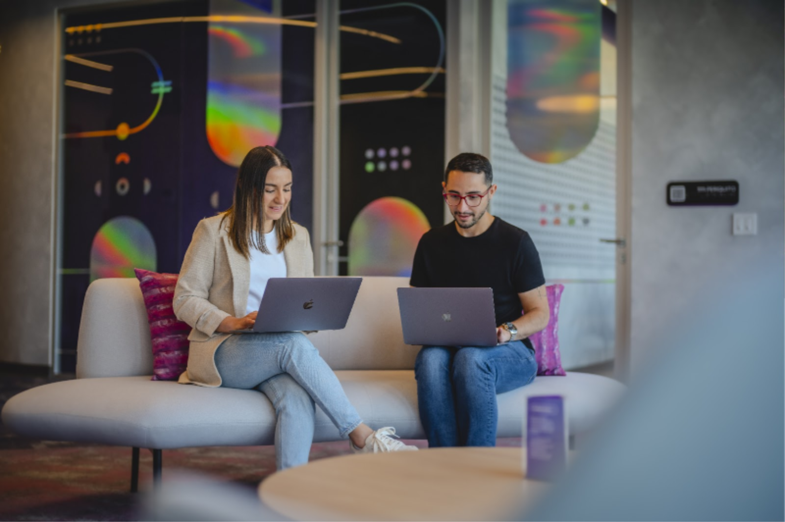 Man and woman with a laptop on a sofa with a low table in front, in the background an internal office area