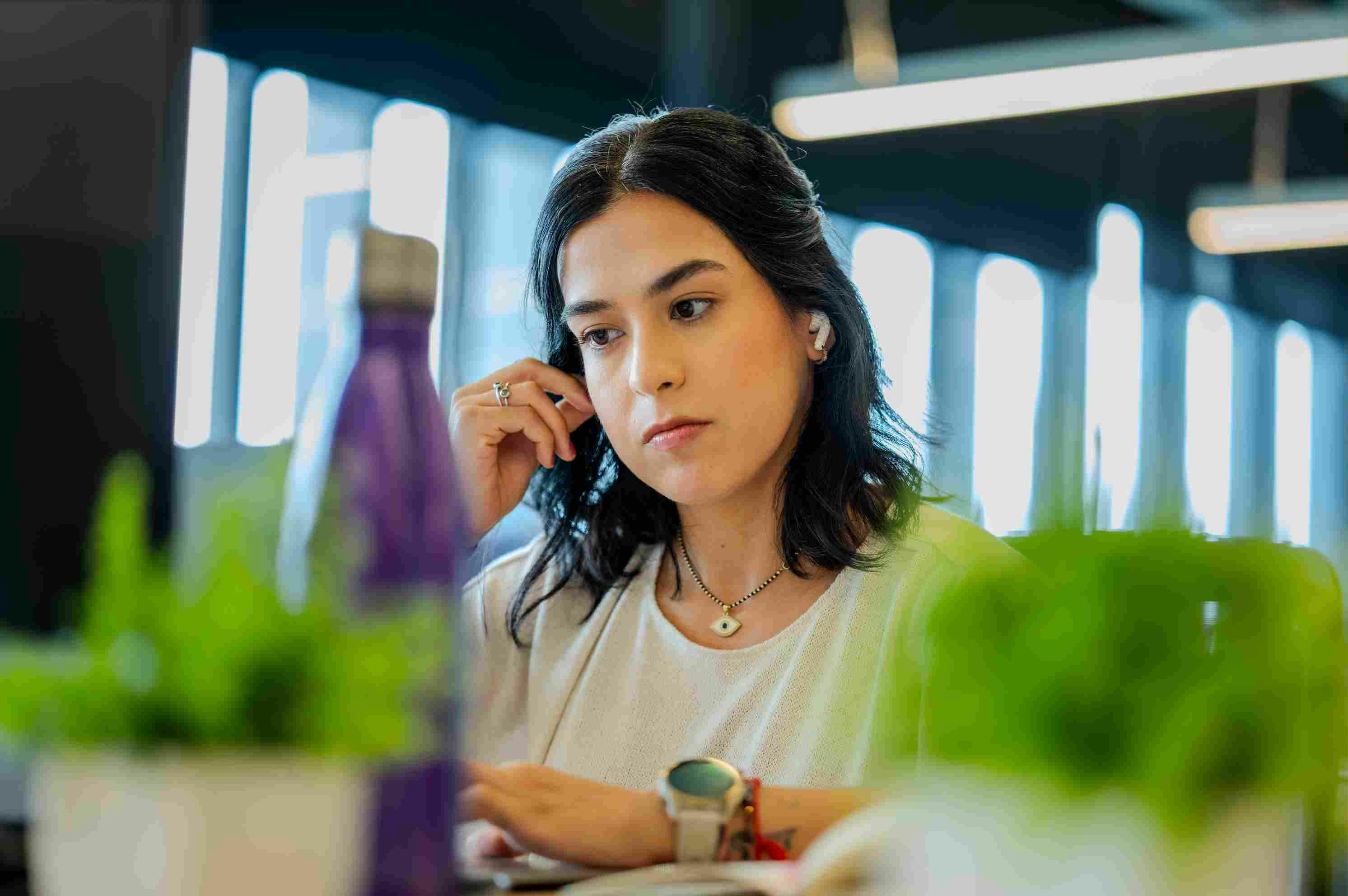 Woman with headphones working focused. In the blurred foreground, there is a purple bottle and a green plant.
