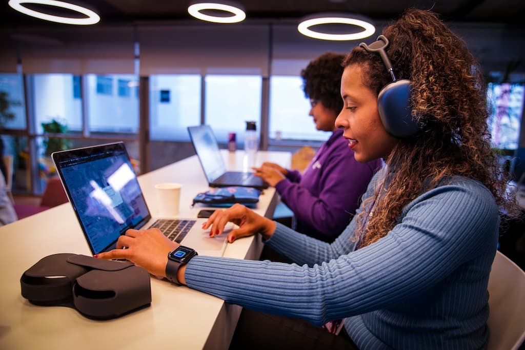A woman wearing a headset uses a laptop at the office, beside colleagues also working.