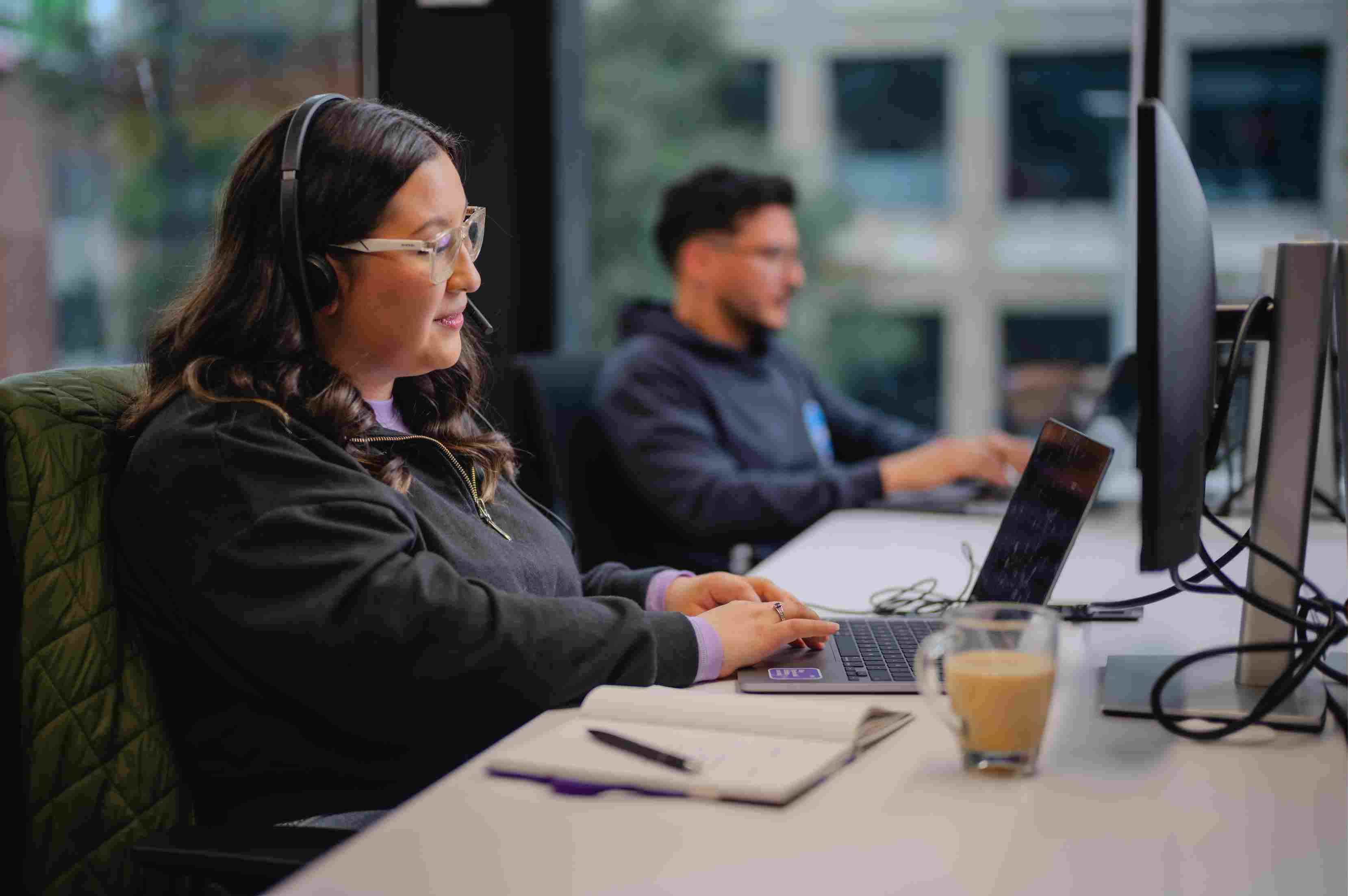 Woman with glasses and a headset working on her notebook with a coffee mug next to her, in a corporate environment.