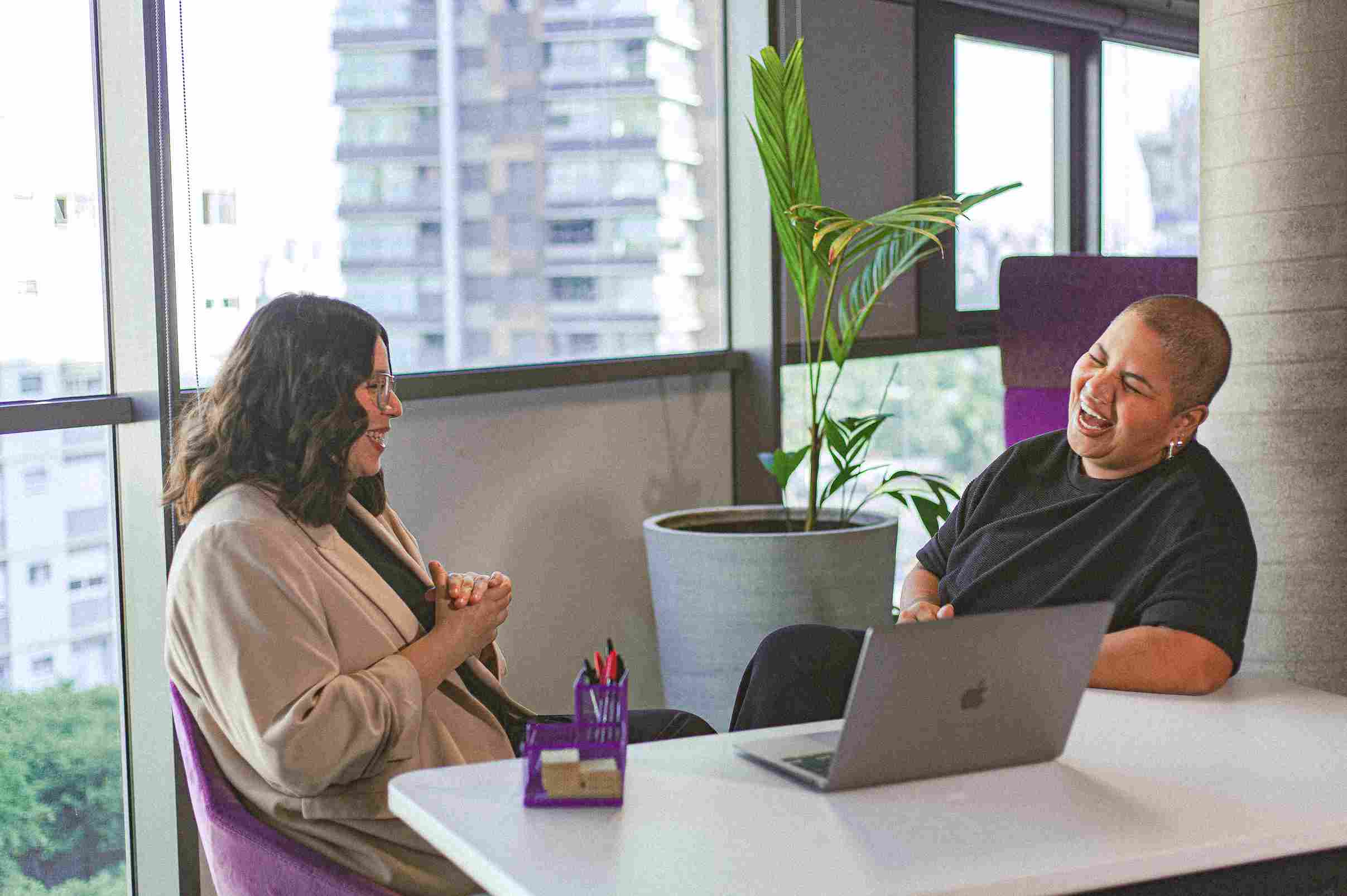 Dos personas sonriendo y conversando animadamente en una mesa con una computadora portátil, cerca de una ventana en la oficina.