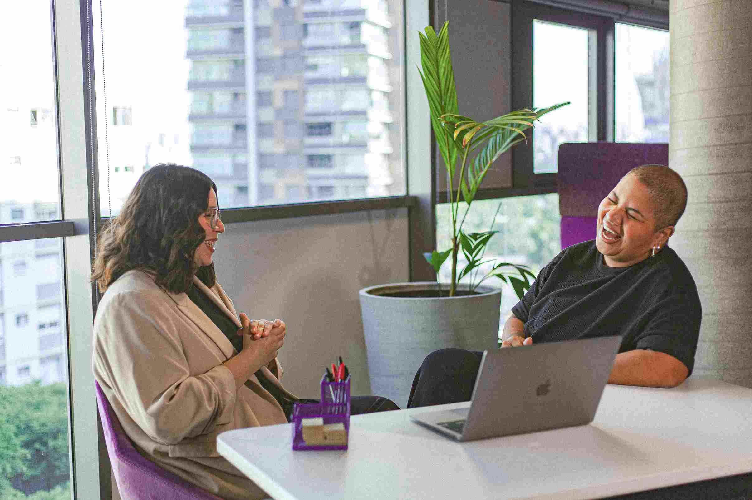 Two people smiling and talking excitedly at a table with a laptop, near a window in the office.