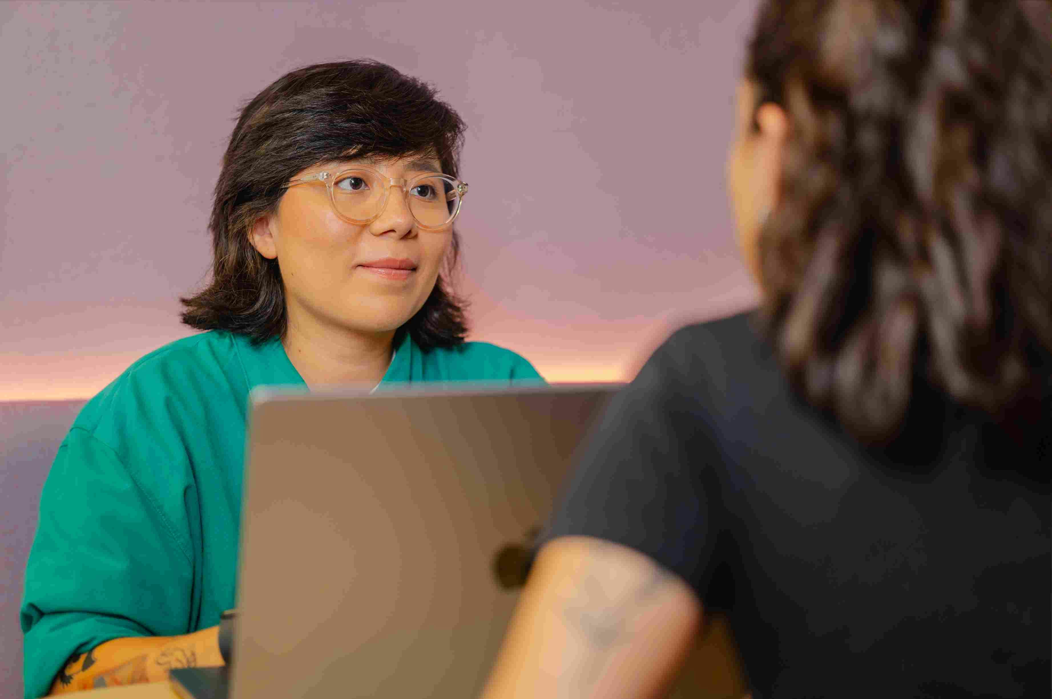 Woman wearing glasses and a green shirt looking attentively at someone, with an open laptop in front of her.
