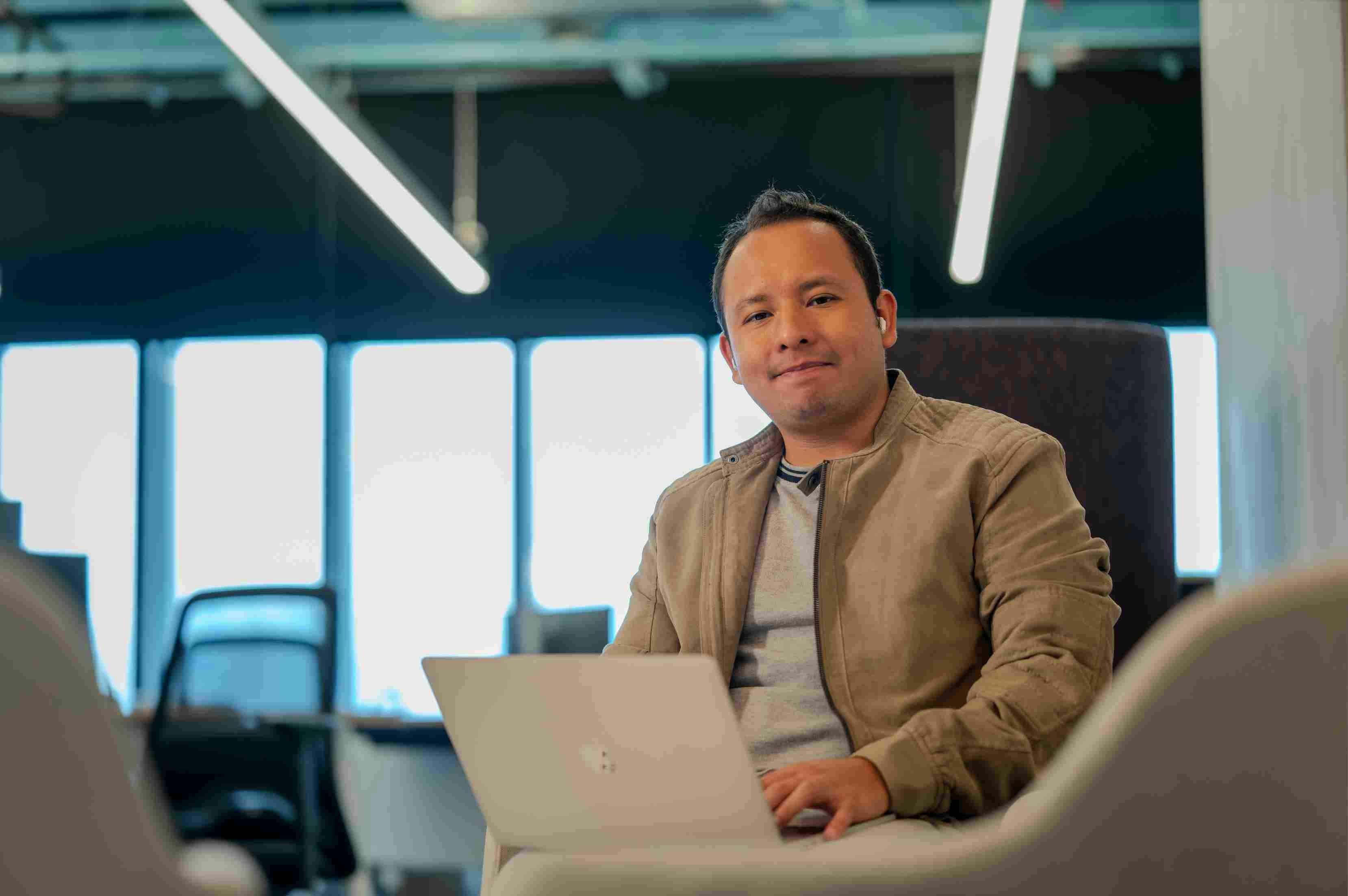 Man smiling at the camera with a laptop on his lap, sitting in a gray chair in a modern office.