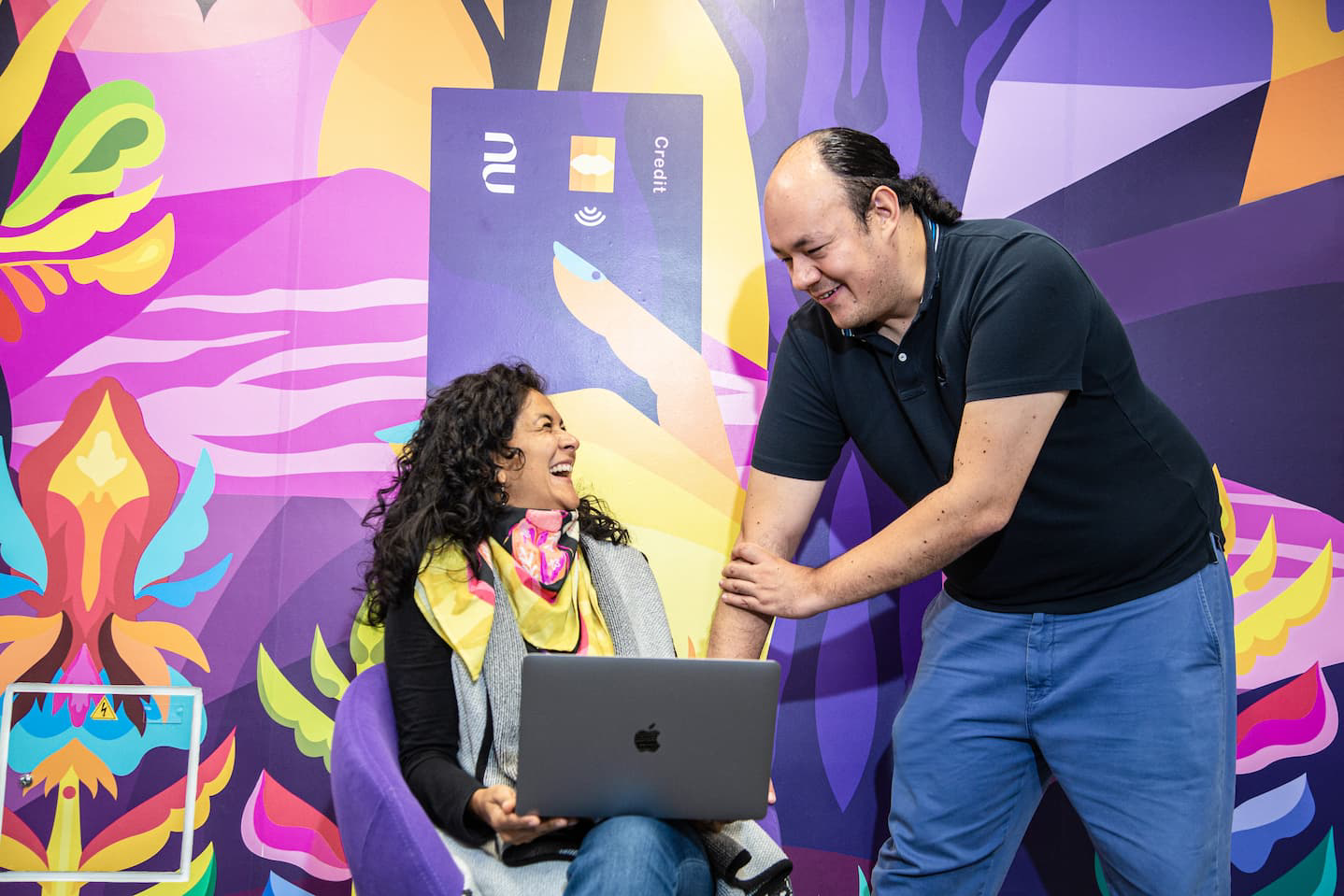 Two people talk in a colorful space while one of them uses a laptop.
