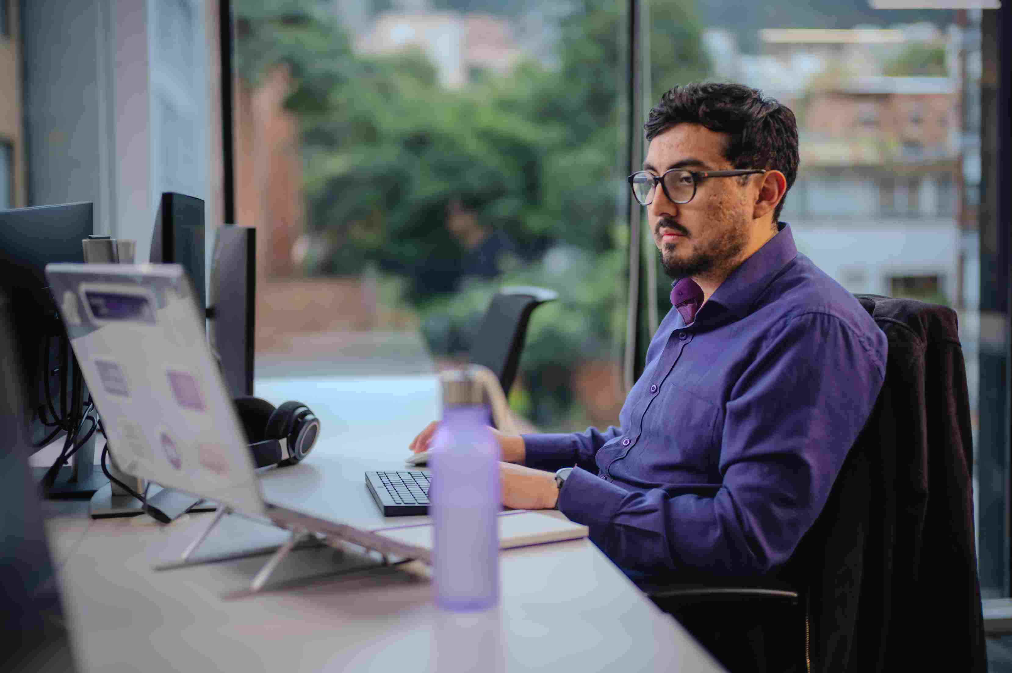 Hombre con gafas y camisa morada trabajando concentrado en un portátil en una mesa de oficina con una botella morada.