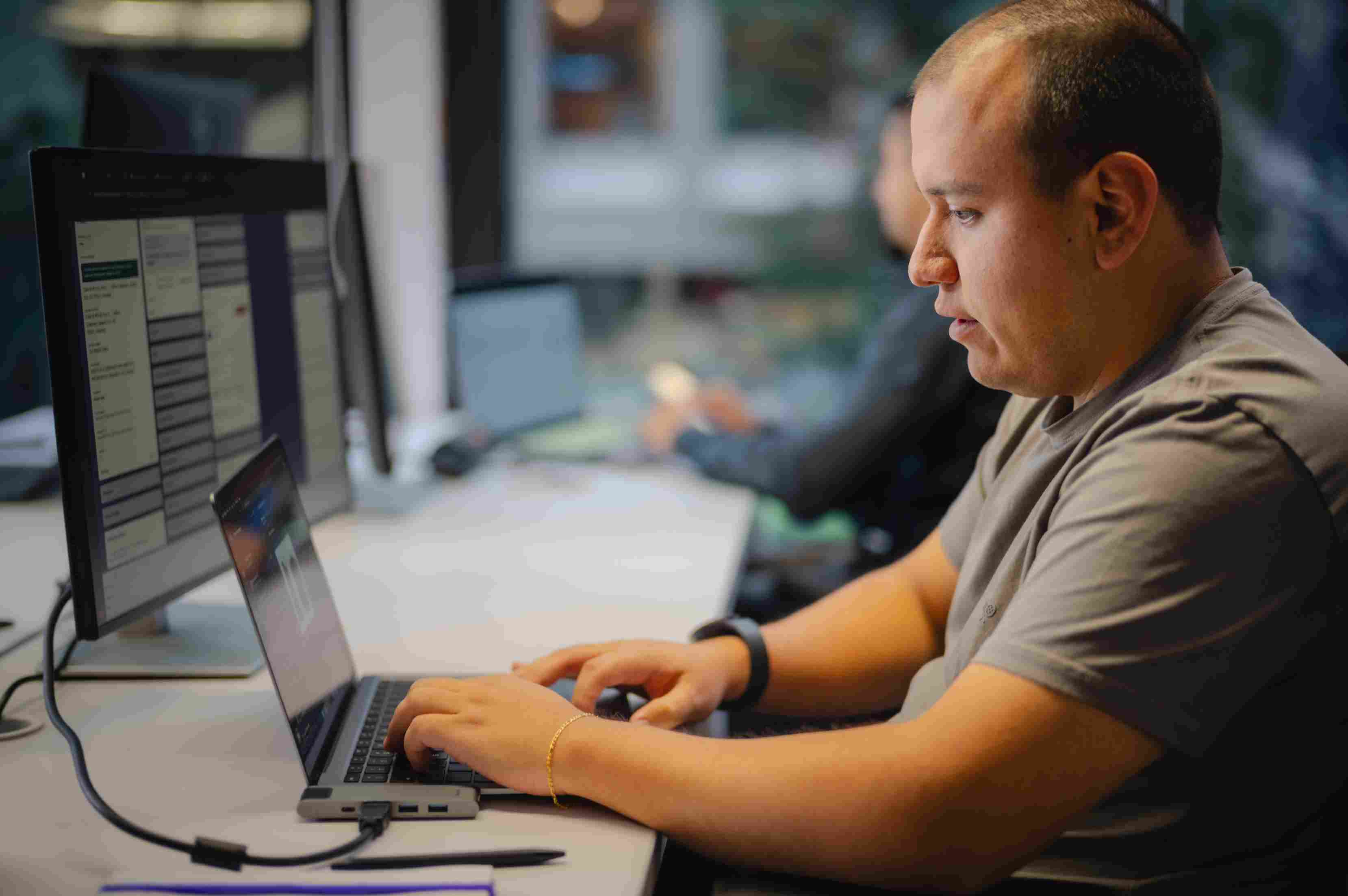 Hombre concentrado escribiendo en un portátil conectado a un monitor grande en un escritorio de oficina.