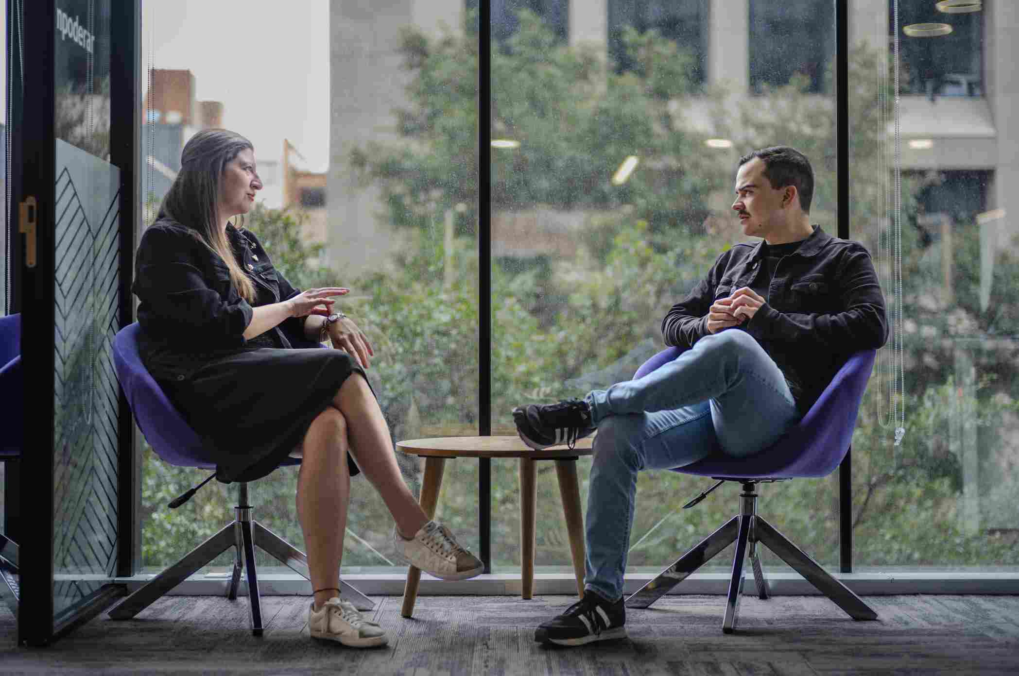Woman and man talking in purple armchairs near a large window overlooking trees.