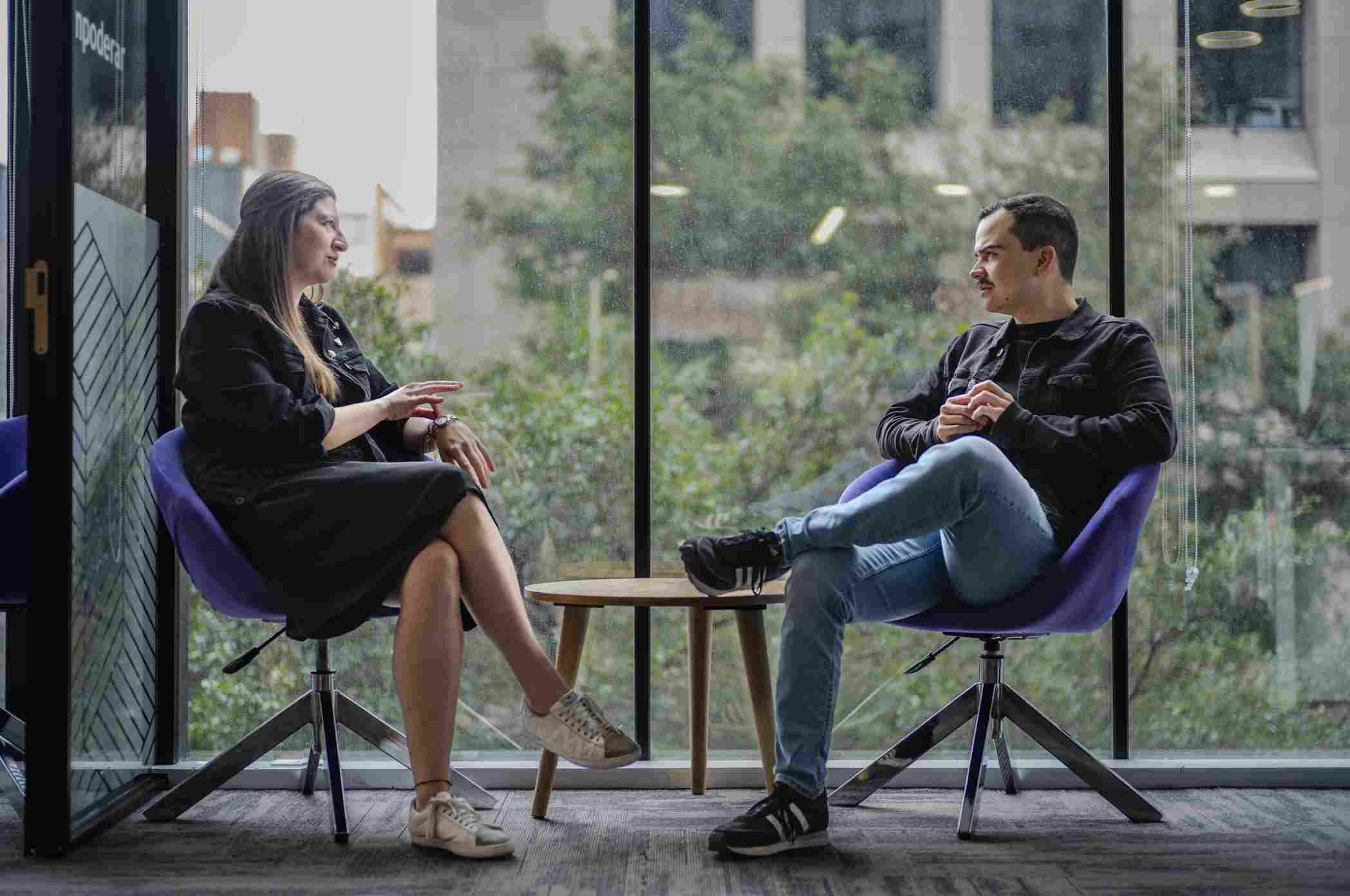 Woman and man talking in purple armchairs near a large window overlooking trees.