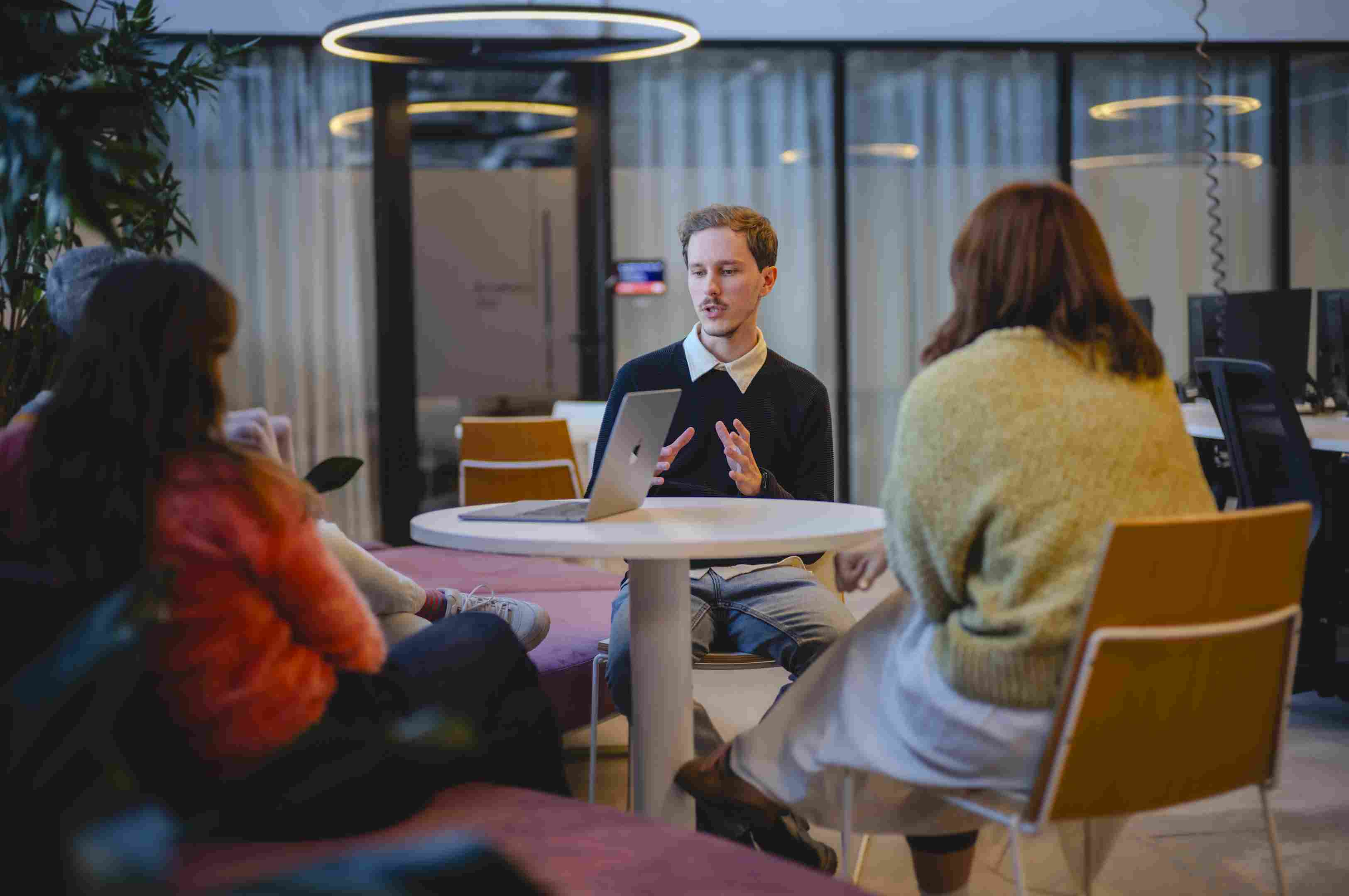 Four people talking at a round table, one of them with a laptop, in the background glass walls with white curtains