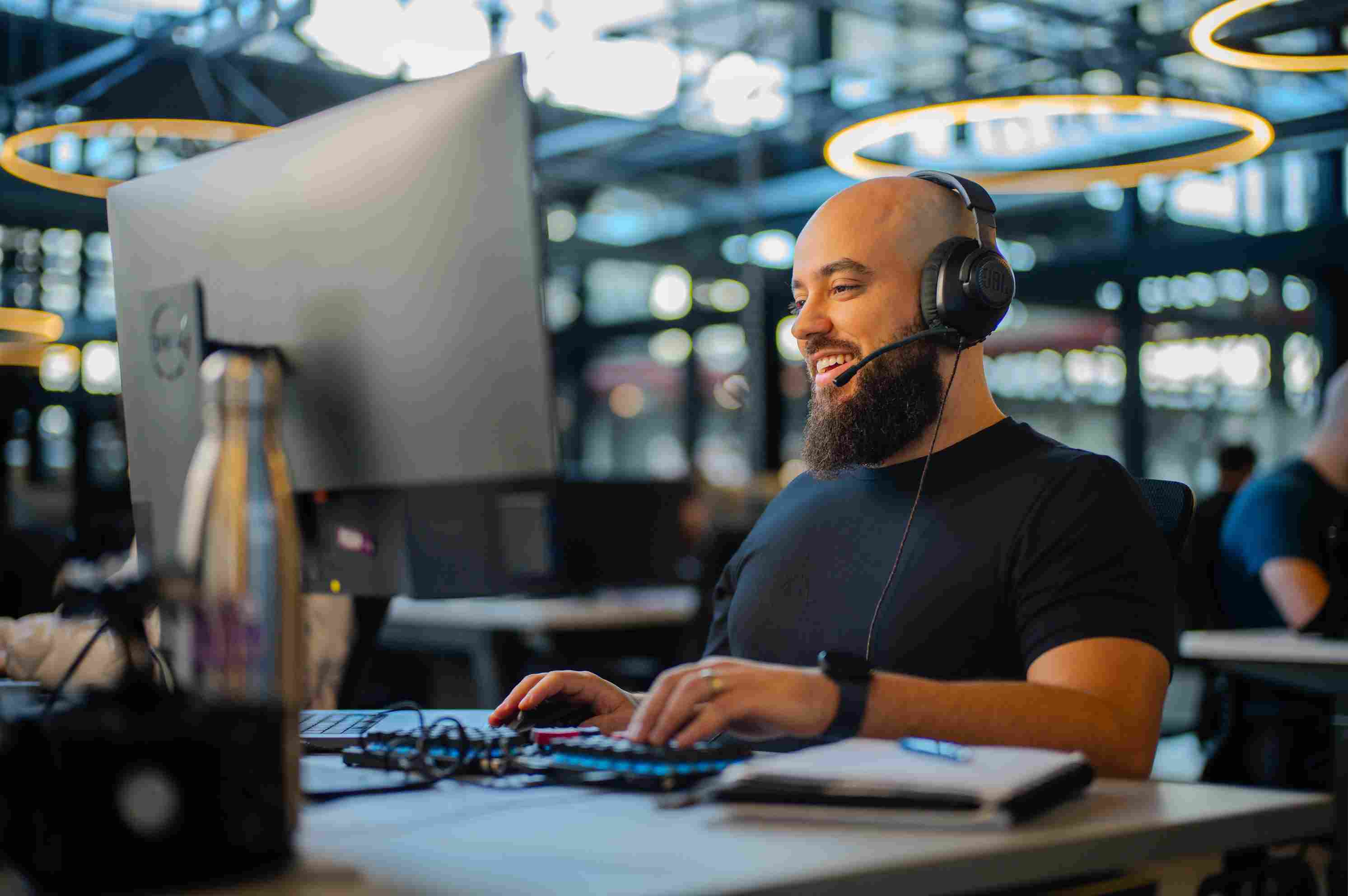 Hombre calvo con barba usando auriculares, sonriendo mientras mira el monitor en la oficina.