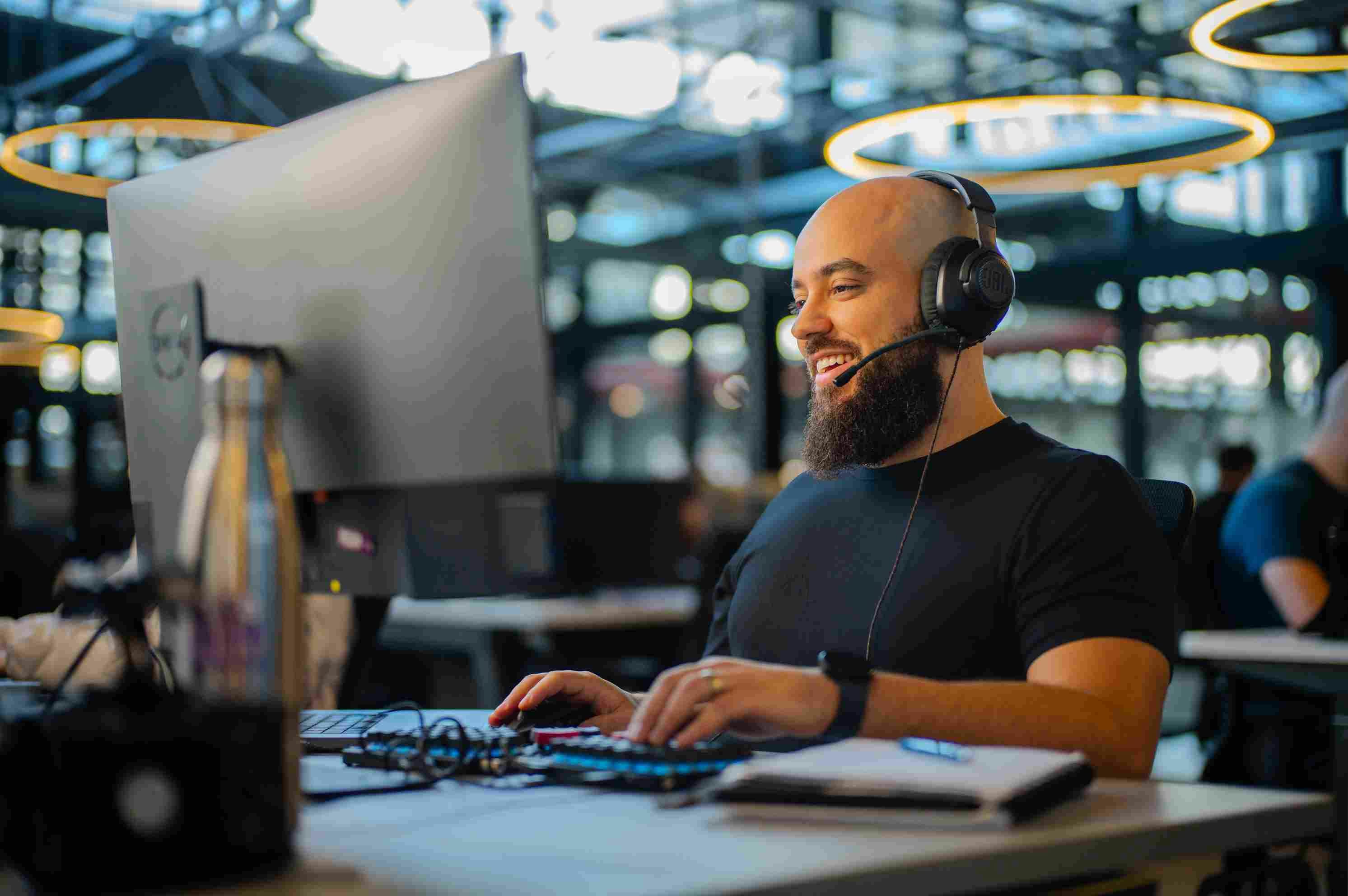 Hombre calvo con barba usando auriculares, sonriendo mientras mira el monitor en la oficina.