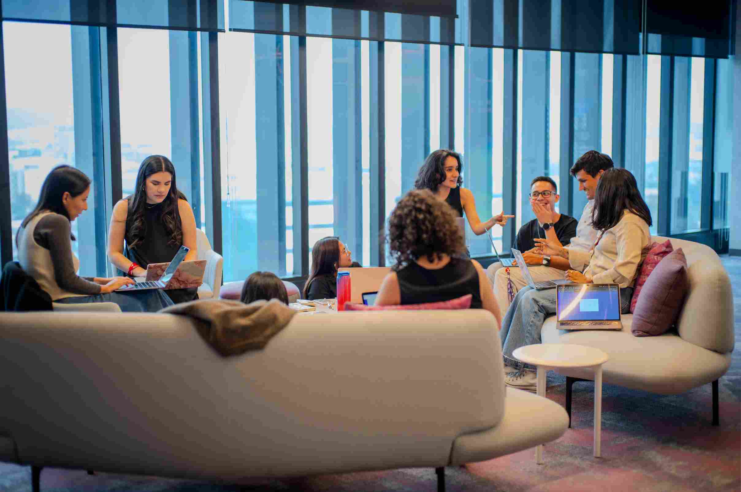 Gran grupo de profesionales conversando y trabajando con portátiles en sofás blancos en una oficina con ventanas.