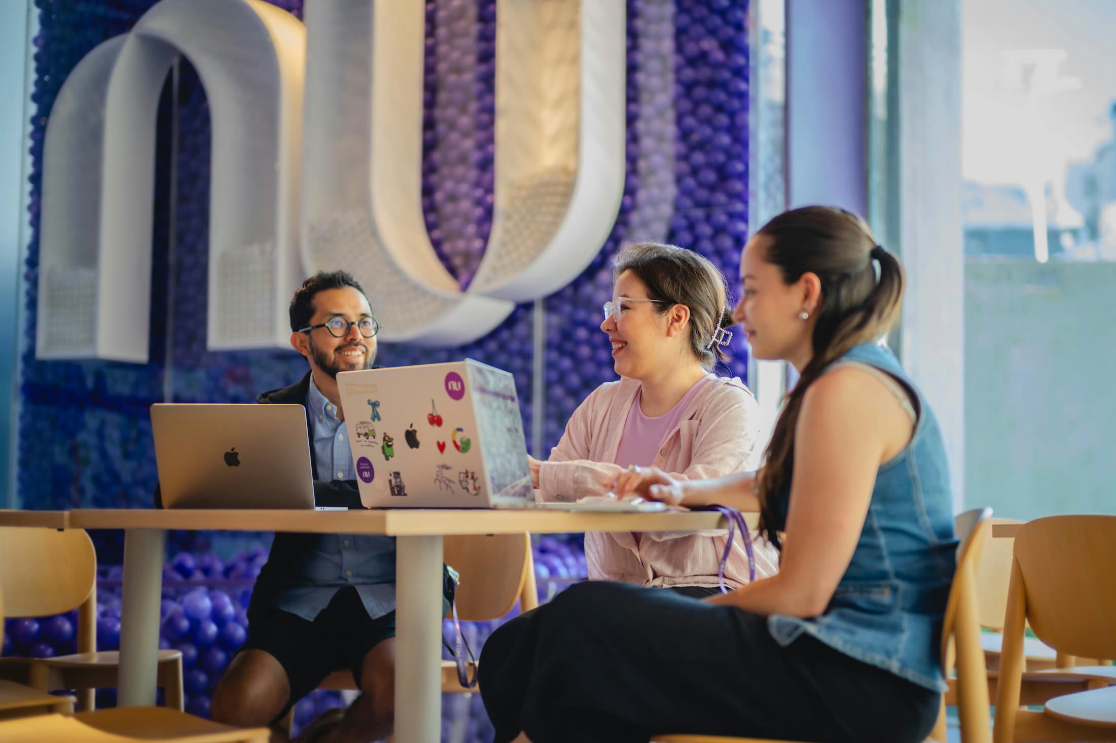 Three people smiling and talking at a round table with a laptop, with a purple wall and the white Nubank symbol in the background