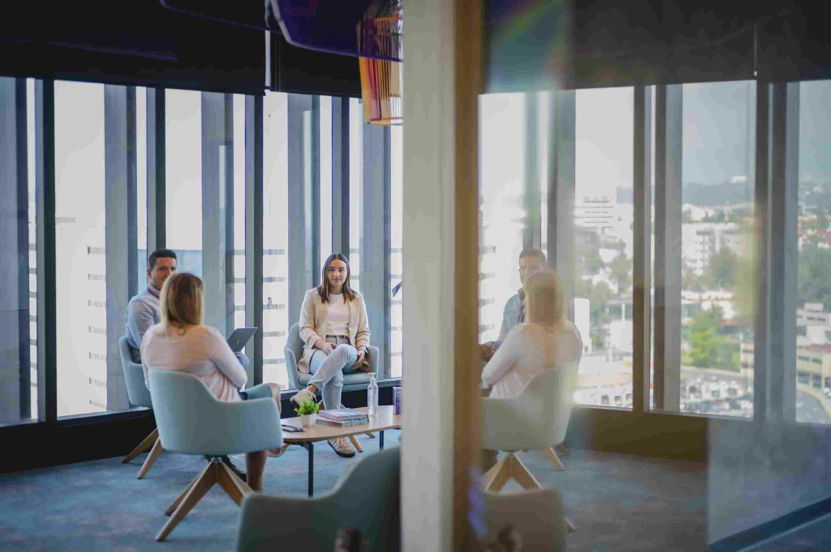 Four people talking while sitting in blue armchairs near a large window with a view of the city.