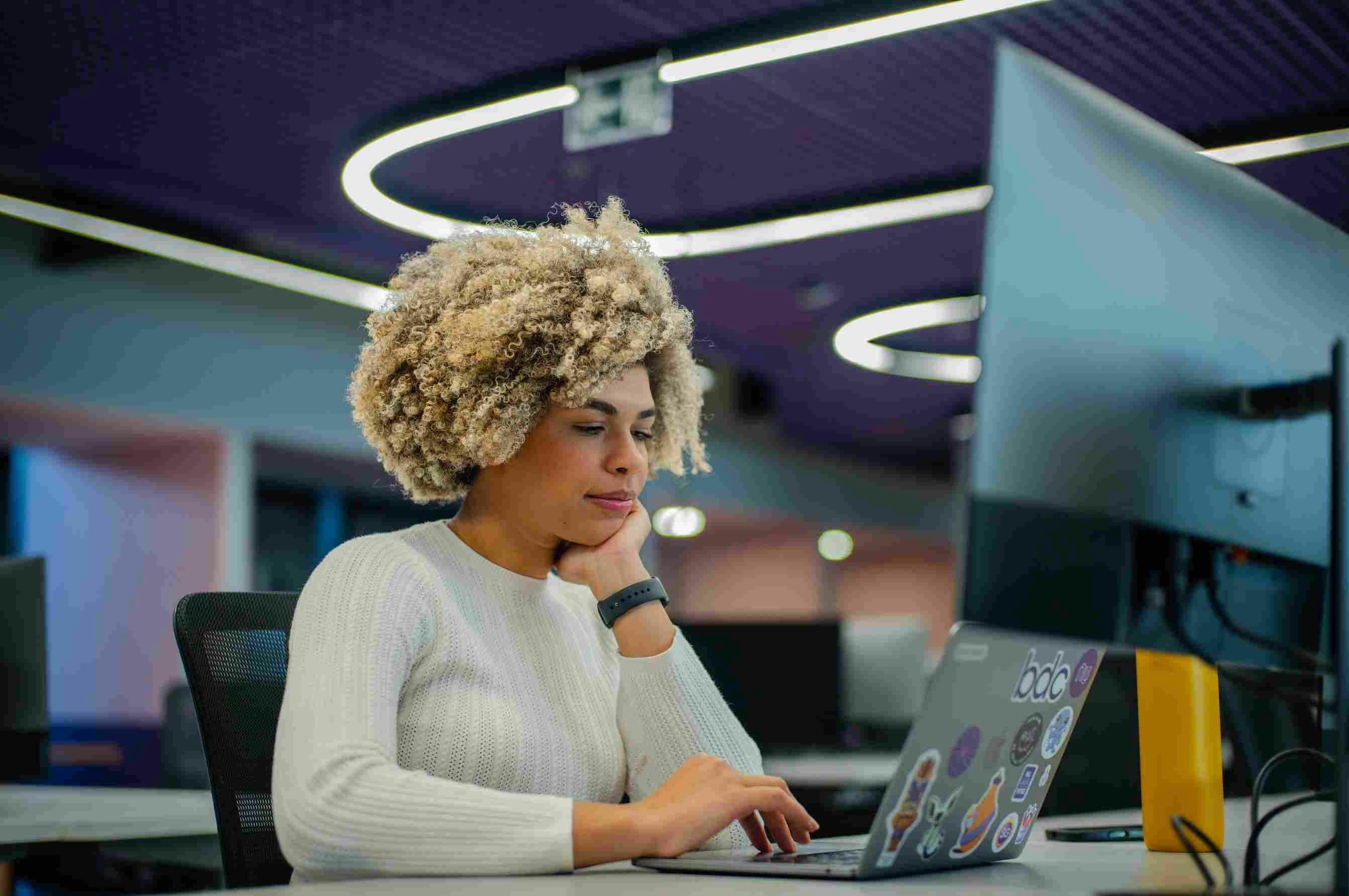 Curly-haired woman working thoughtfully on a notebook with stickers, in an office with lights on the ceiling.