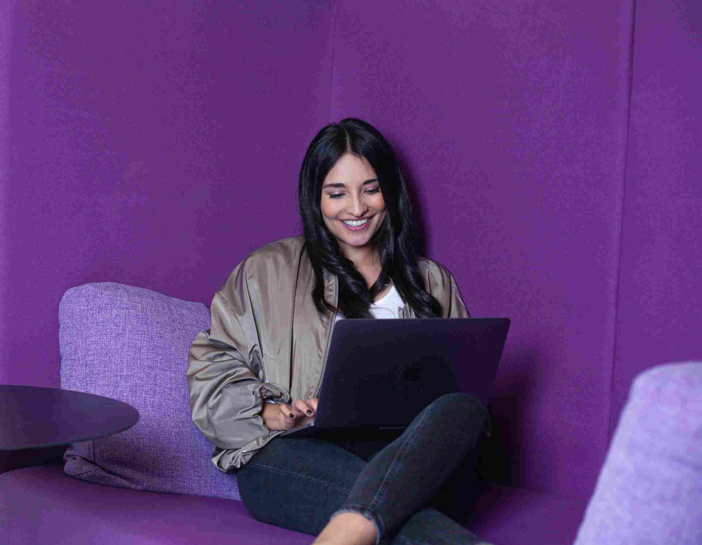 Woman smiling while typing on her laptop in a purple booth.