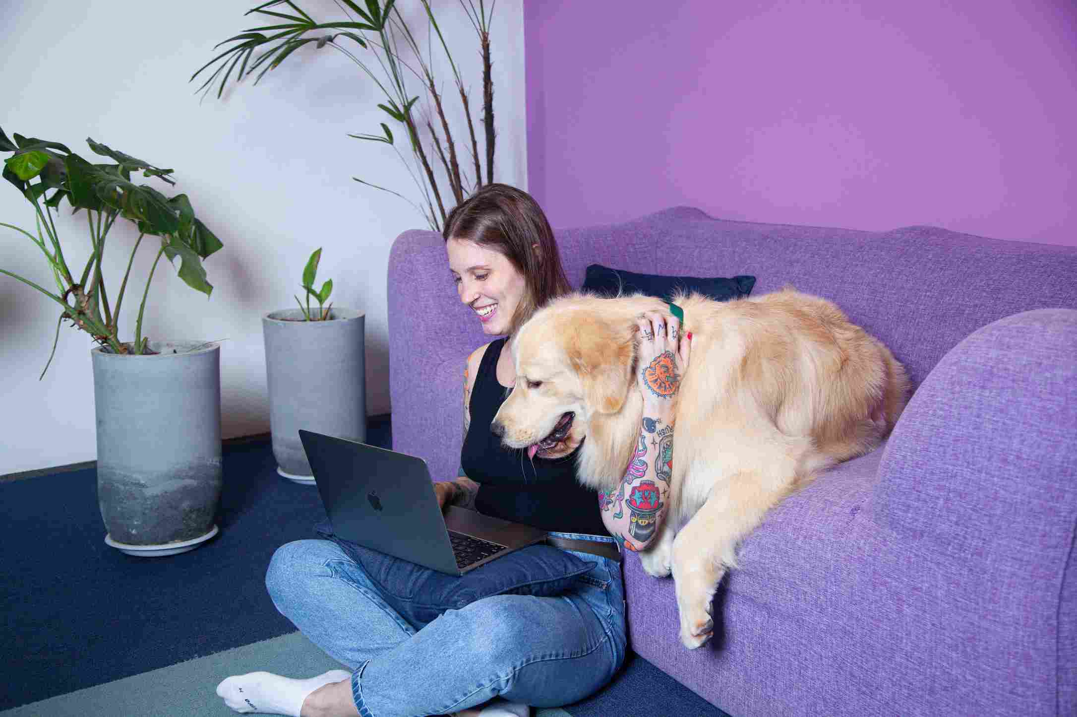 Mujer usa su laptop en el suelo junto a un perro en el sofá.