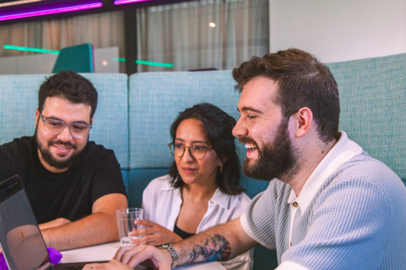 Three coworkers smiling while looking at a laptop screen in an office booth.