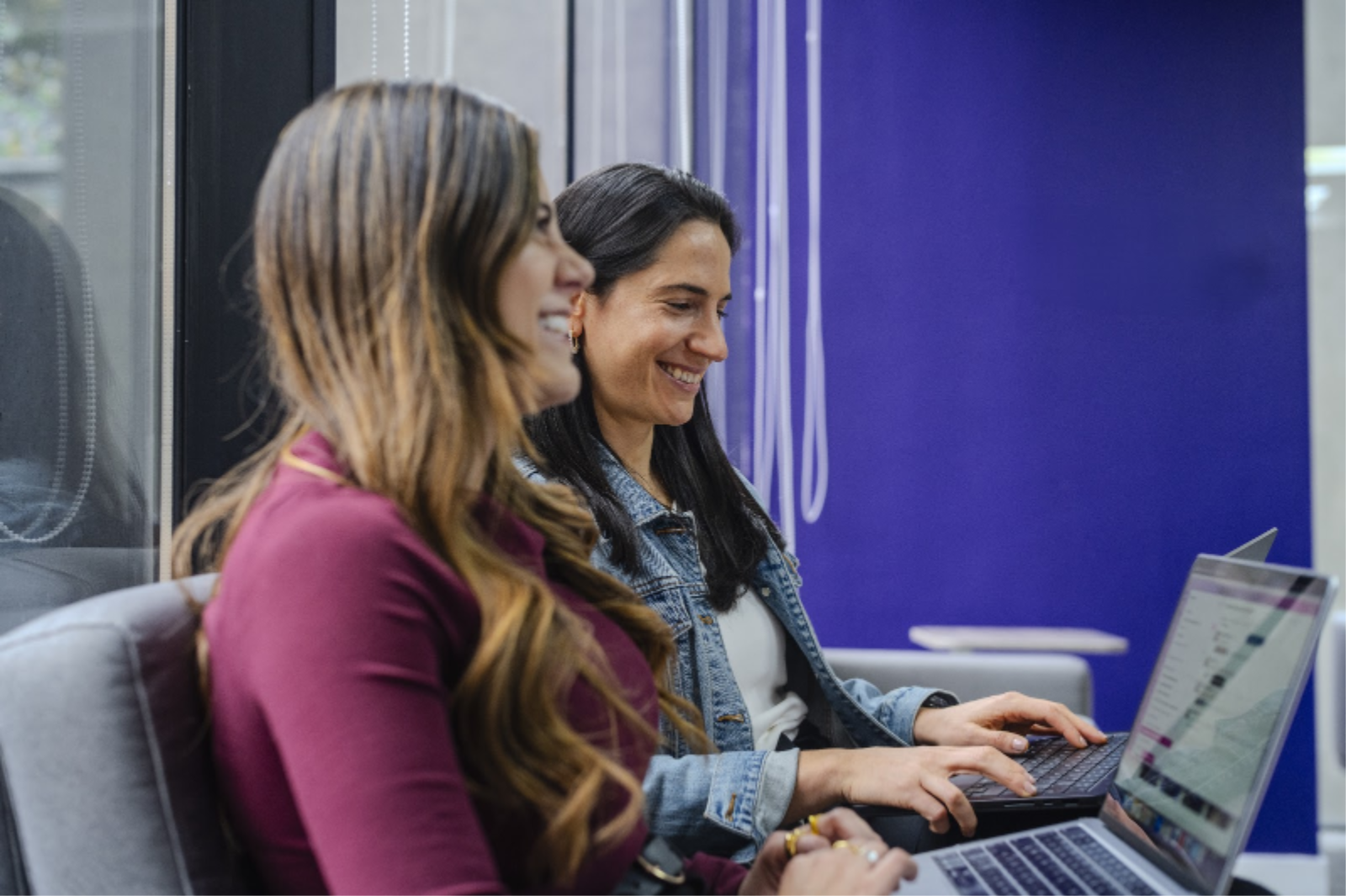Dos mujeres, de lado, sonriendo usando la computadora portátil, al fondo una pared morada y un ventanal de vidrio.