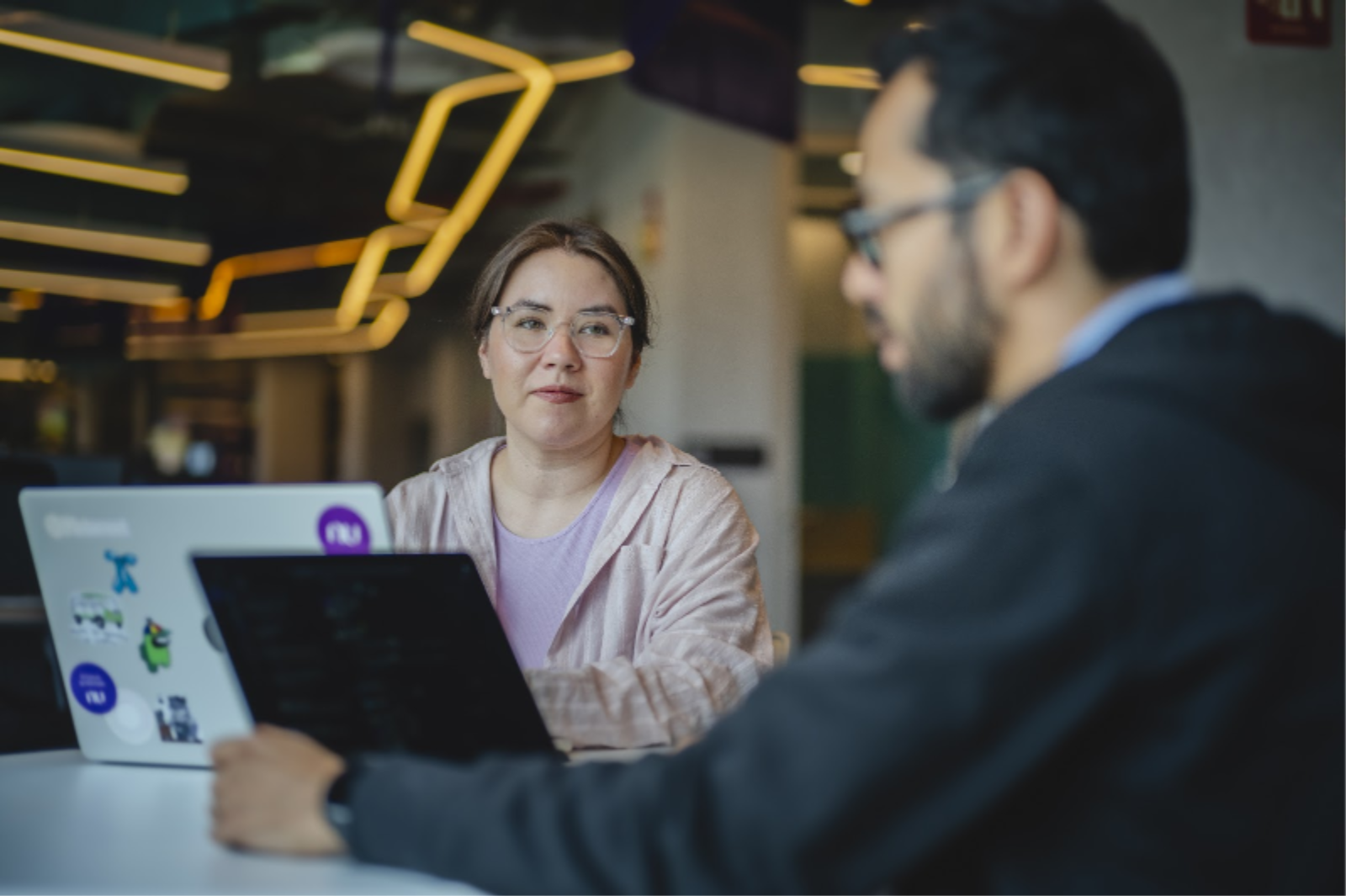 Woman in a lilac t-shirt, man with a beard, sitting together at a table, with the office interior in the background.
