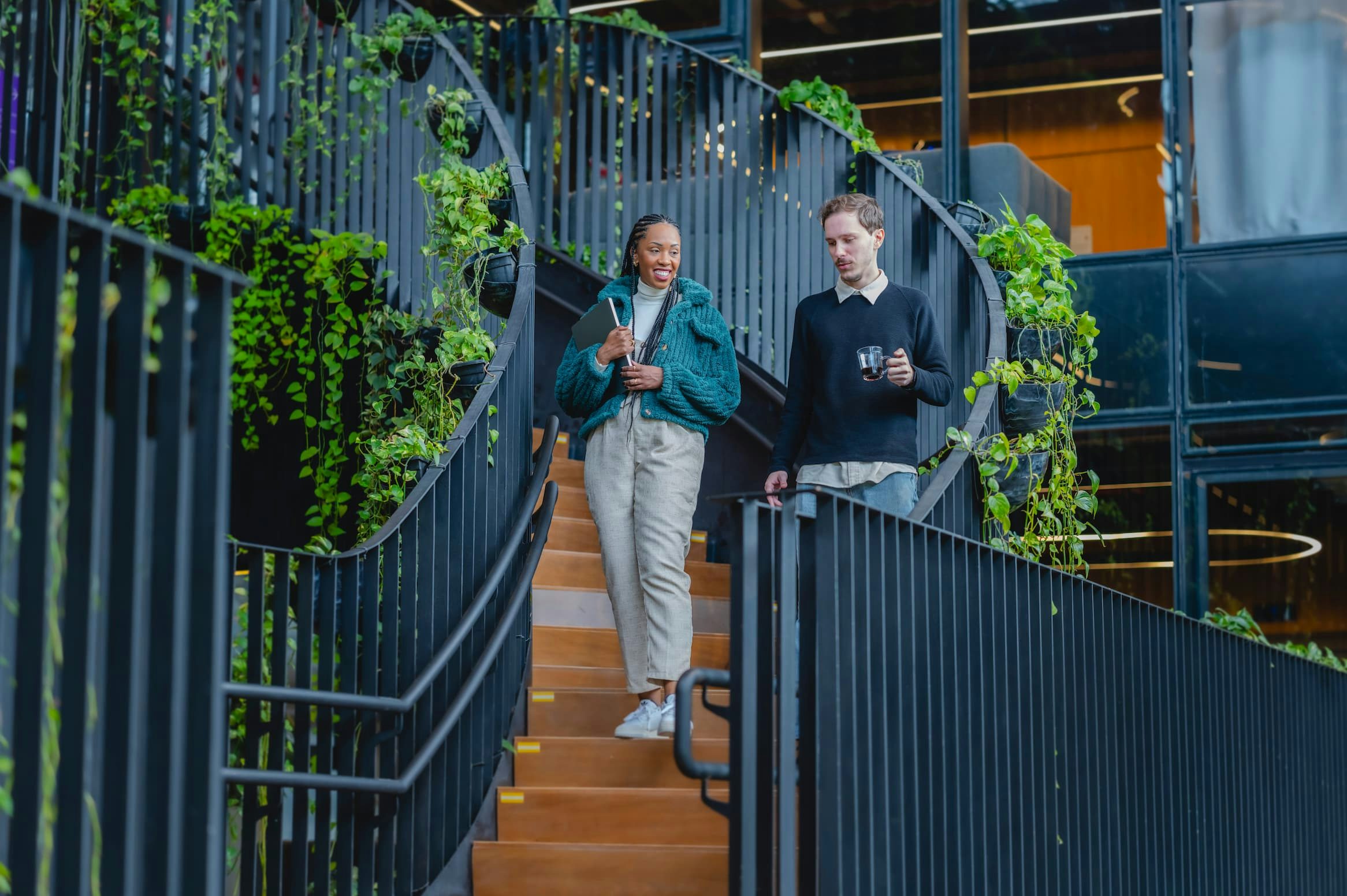 A woman in a green coat and a man in a black sweater descending a yellow-and-black staircase surrounded by plants.