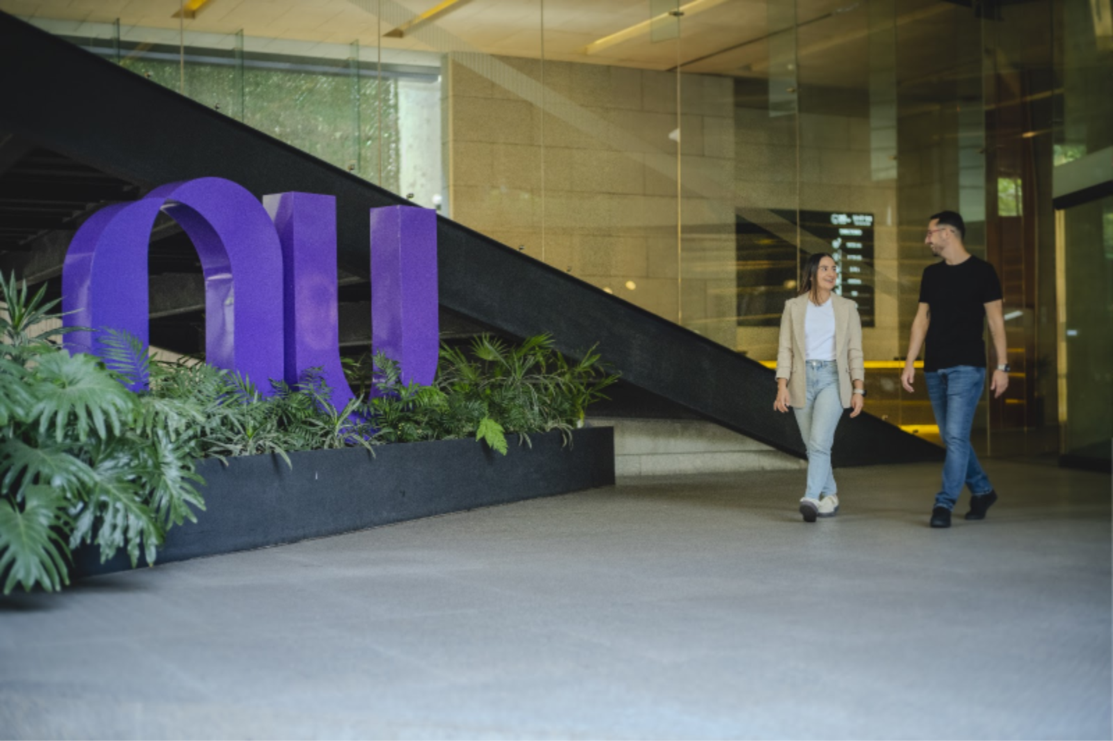 Hombre y mujer caminan por el vestíbulo de una oficina junto a un gran logo púrpura de Nubank rodeado de plantas.