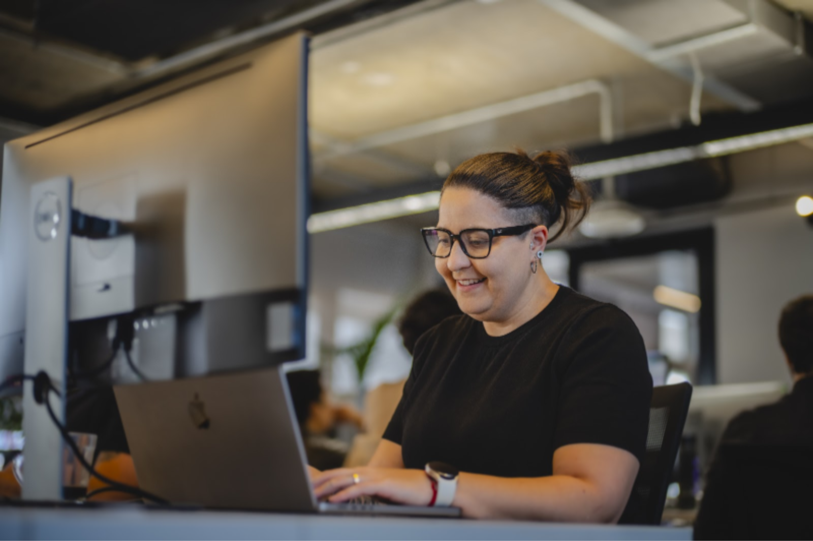 Persona con camiseta negra y gafas, sentada en un escritorio trabajando en una computadora portátil y con una pantalla de apoyo