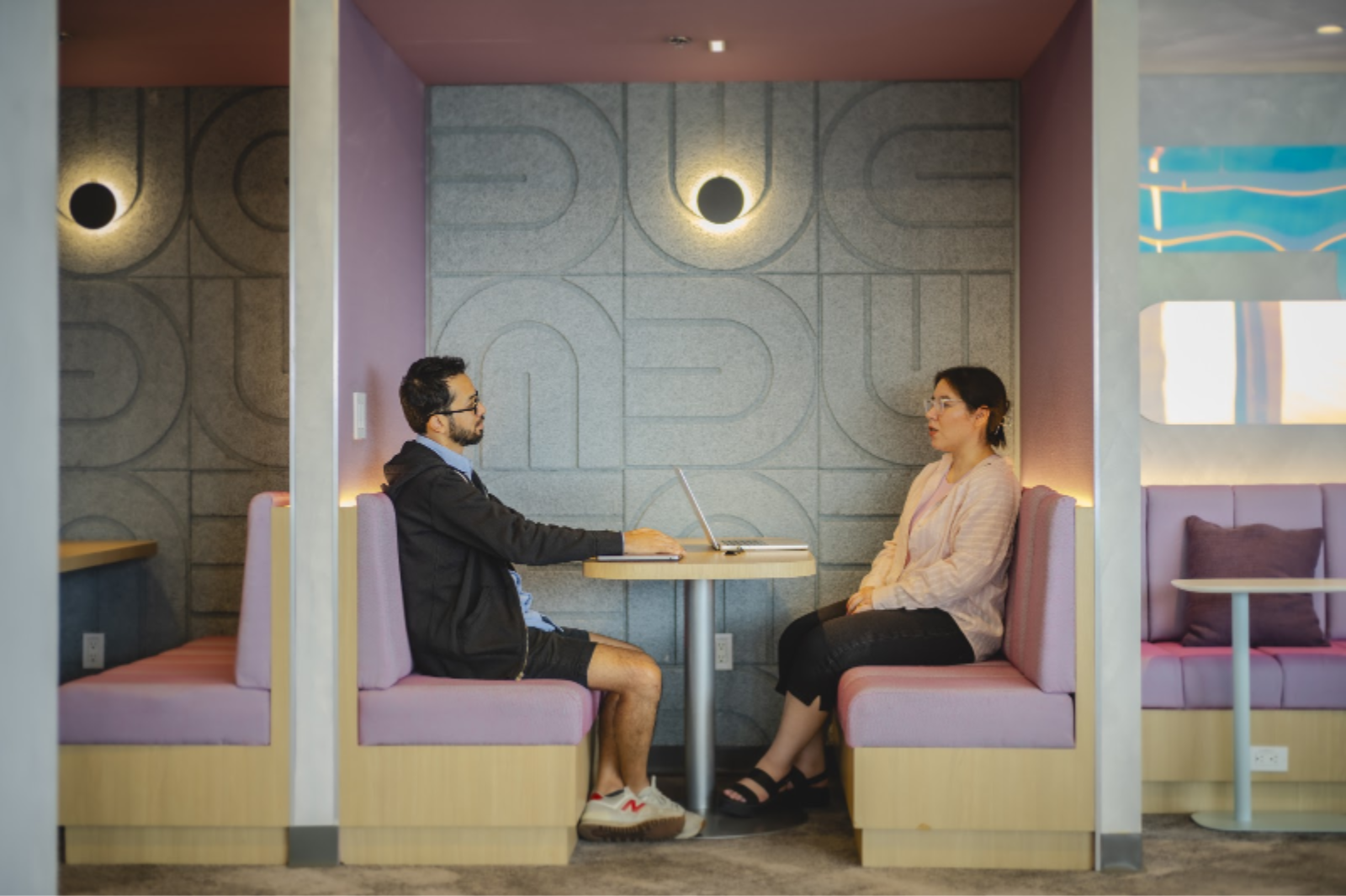 Hombre y mujer conversando en una cabina de oficina con sofás morados y pared con textura ovalada.