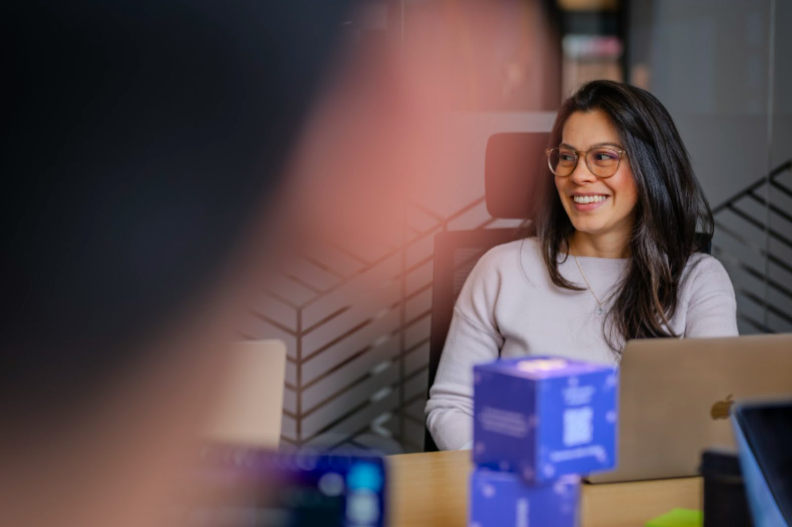 Woman with glasses smiling at her desk with a laptop and blue decorative Nubank cubes.