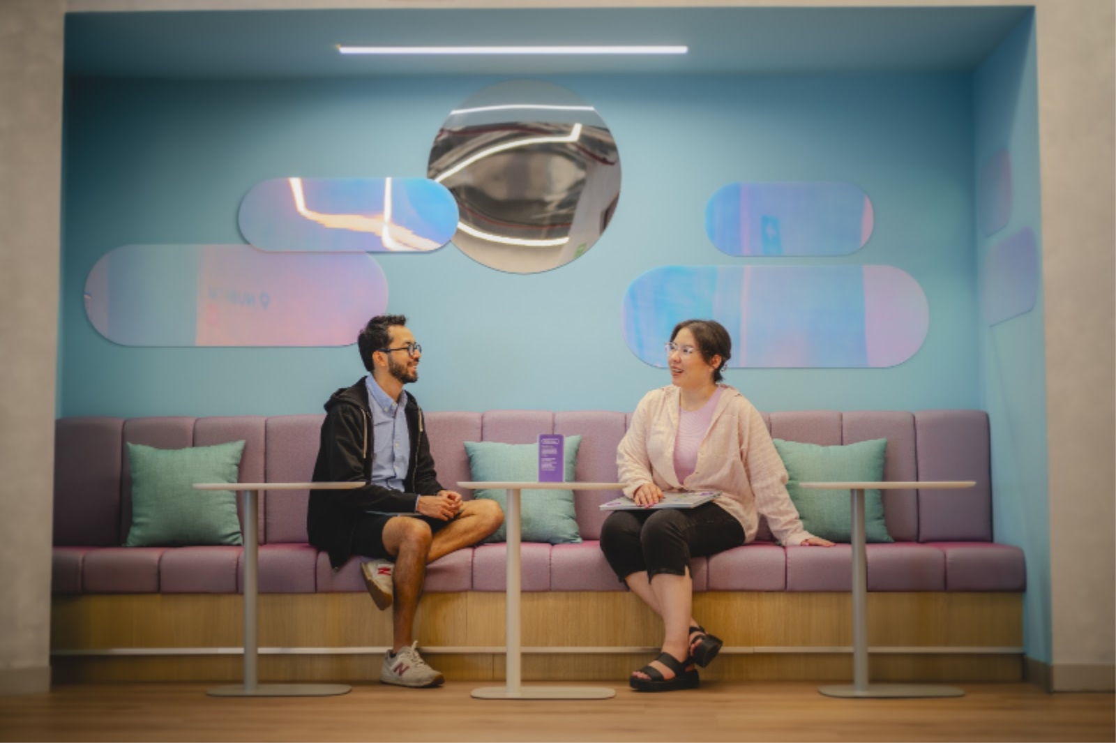 Man and woman talking on a lilac sofa with small tables in front, light blue wall in the background, and wooden floor.