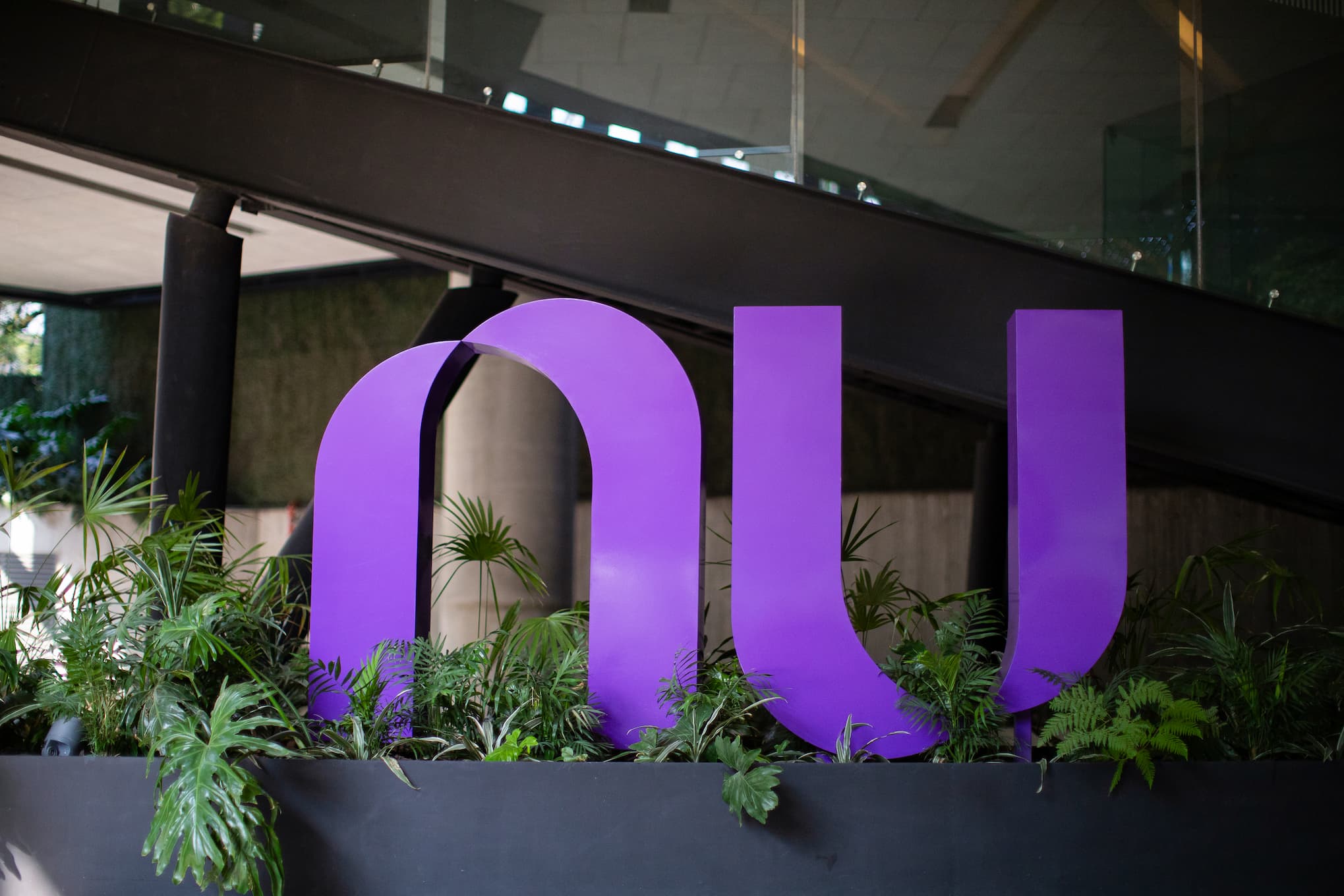 Close no grande logotipo roxo do Nu instalado em um canteiro com plantas na entrada de um edifício moderno.