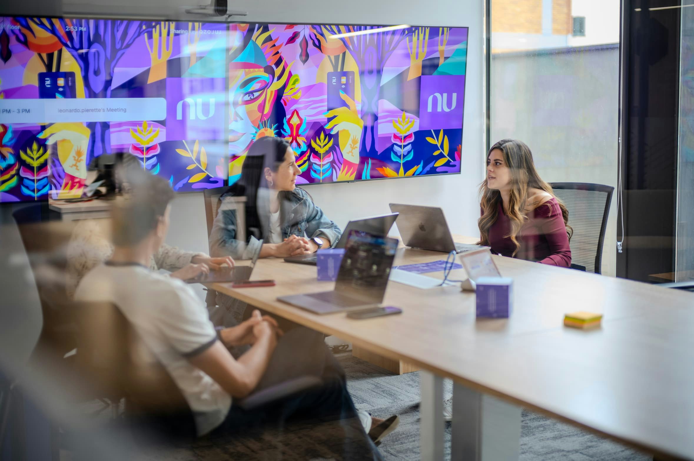People seen through a glass wall, sitting at a table and chatting in a meeting room; in the background, a display board with the Nubank logo.