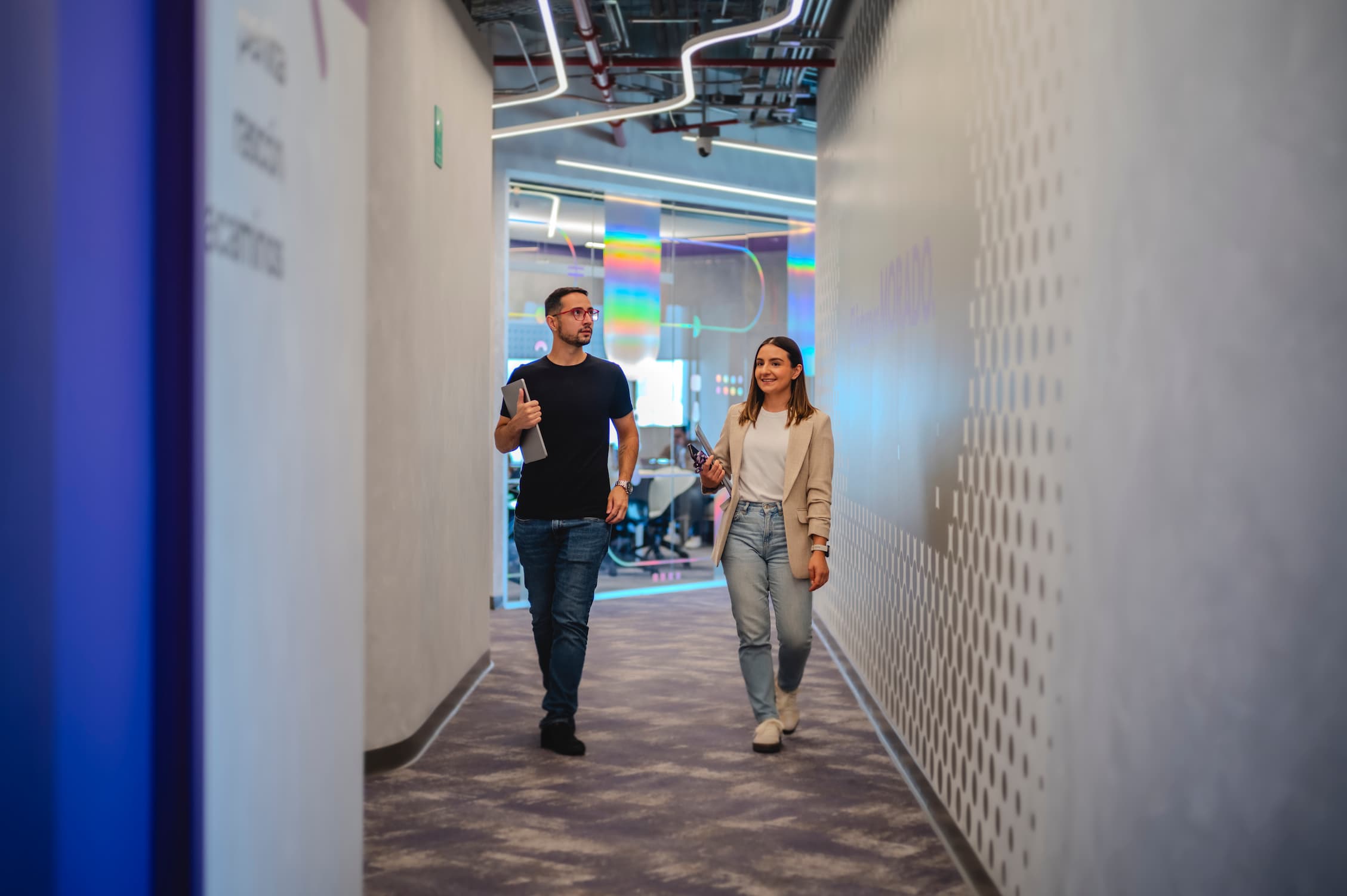 A man in a black T-shirt and jeans and a woman in a white T-shirt, a beige coat, and jeans chatting in a hallway.