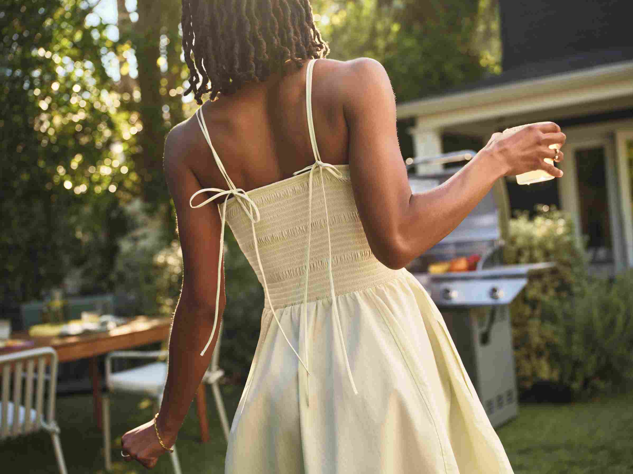 Mulher de costas com vestido de verão e copo na mão, em um quintal ensolarado com uma churrasqueira ao fundo.