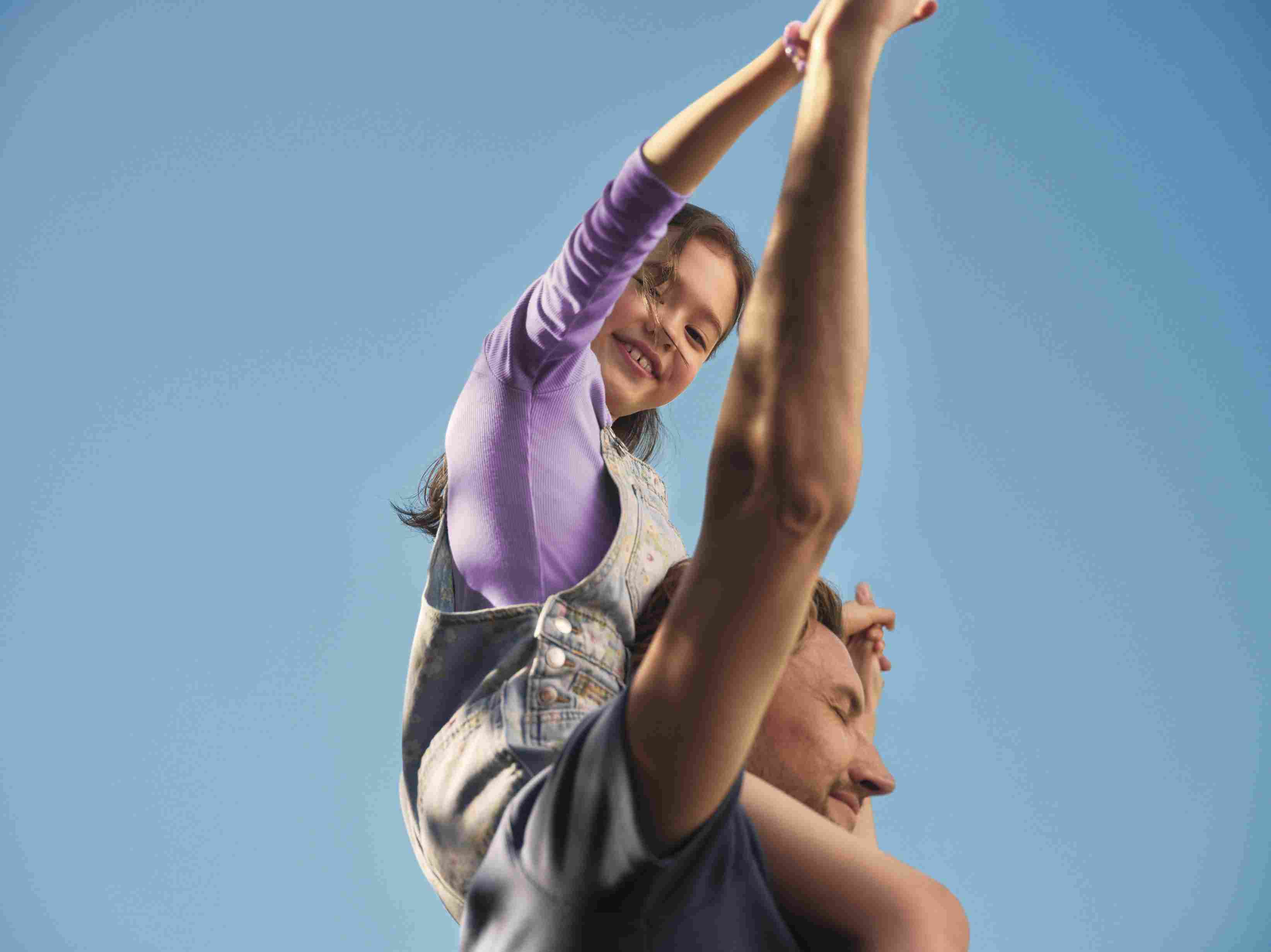 Menina sorridente sentada nos ombros de um homem com o céu azul ao fundo, em um momento de diversão em família.