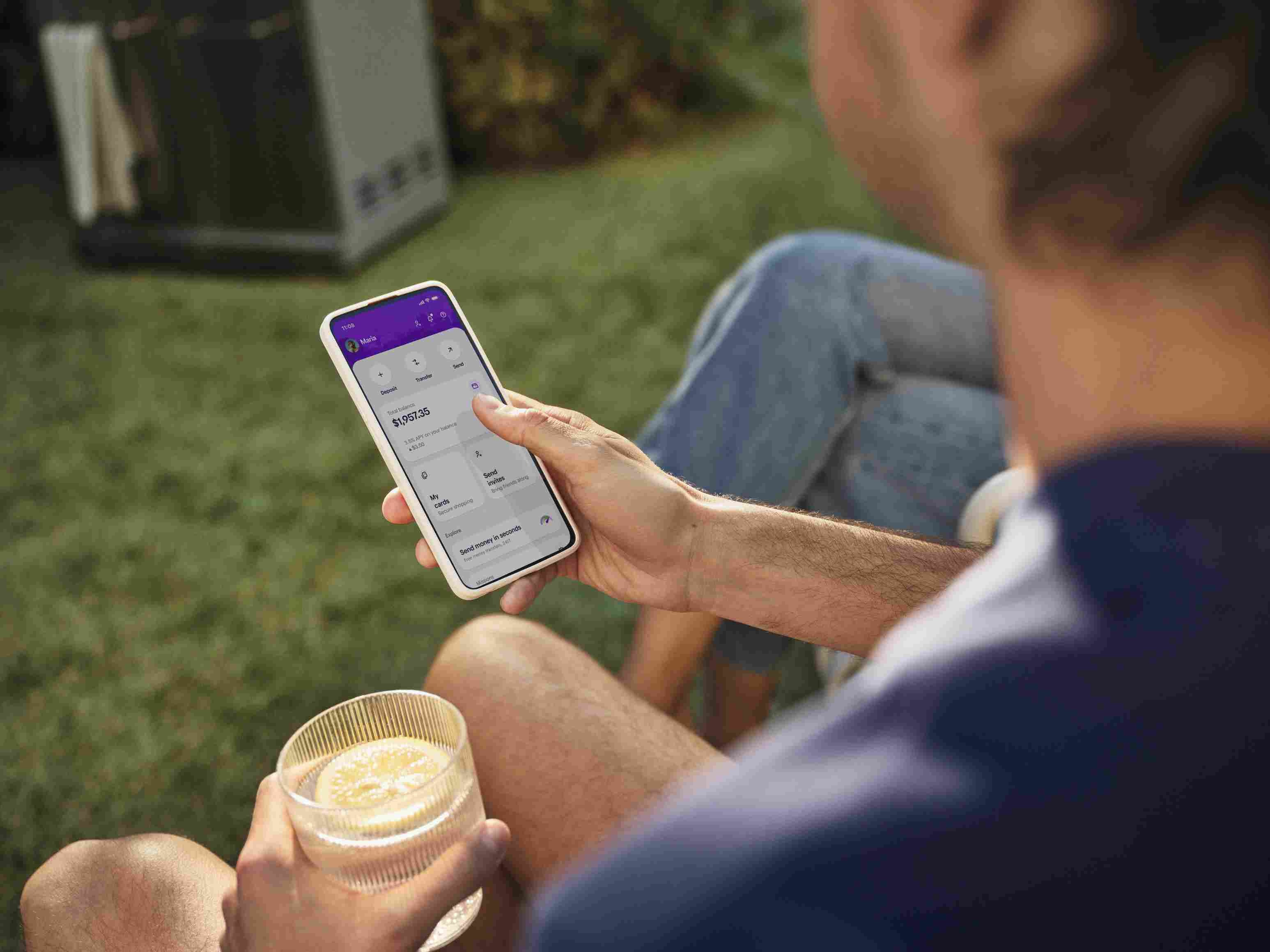 Homem segurando um copo e usando o aplicativo do Nubank no celular. Ao fundo, vê-se a grama e uma churrasqueira.