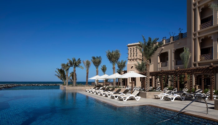 The Sheraton Sharjah Beach Resort & Spa A luxurious poolside view at a resort with white lounge chairs, palm trees, and a clear blue sky overlooking the ocean.