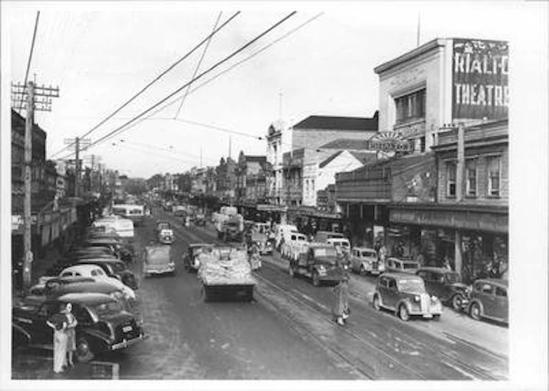Black and white street scene showing cars parked on diagonals on the street. You can see the side of a building painted with a sign for Rialto Theatre