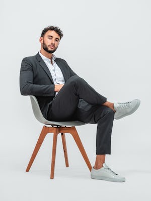 Man wearing Velocity Pant Dark Charcoal, Hybrid Button Down Grey Stripe and Velocity Blazer Dark Charcoal with grey sneakers and sitting in a grey chair