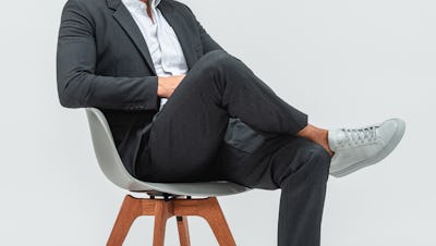 Man wearing Velocity Pant Dark Charcoal, Hybrid Button Down Grey Stripe and Velocity Blazer Dark Charcoal with grey sneakers and sitting in a grey chair