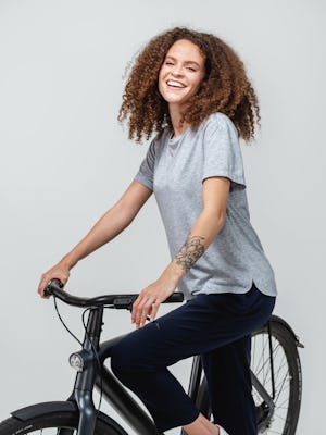 model wearing pale grey heather composite merino boxy tee and navy swift drape pant standing next to a bicycle with hands on handlebars