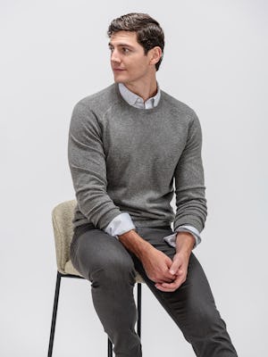 model wearing men's dark grey chroma denim and grey heather stripe hybrid button-down and medium grey atlas merino crew neck sweater sitting in chair with sleeves pulled up 