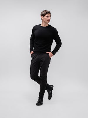 model wearing men's black kinetic tapered pant and black atlas merino crew neck sweater facing forward with legs crossed and hands in pockets
