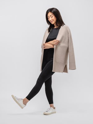 model wearing women's oatmeal composite merino cardigan and black composite merino boxy tee and black joule active legging facing to the side with arms crossed and cardigan draped over shoulders while kicking the air