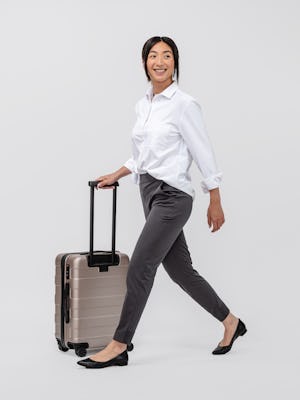 model wearing women's charcoal kinetic pull on pant and white aero zero oversized shirt walking to the side with shirt tucked, sleeves rolled and rolling a travel suitcase